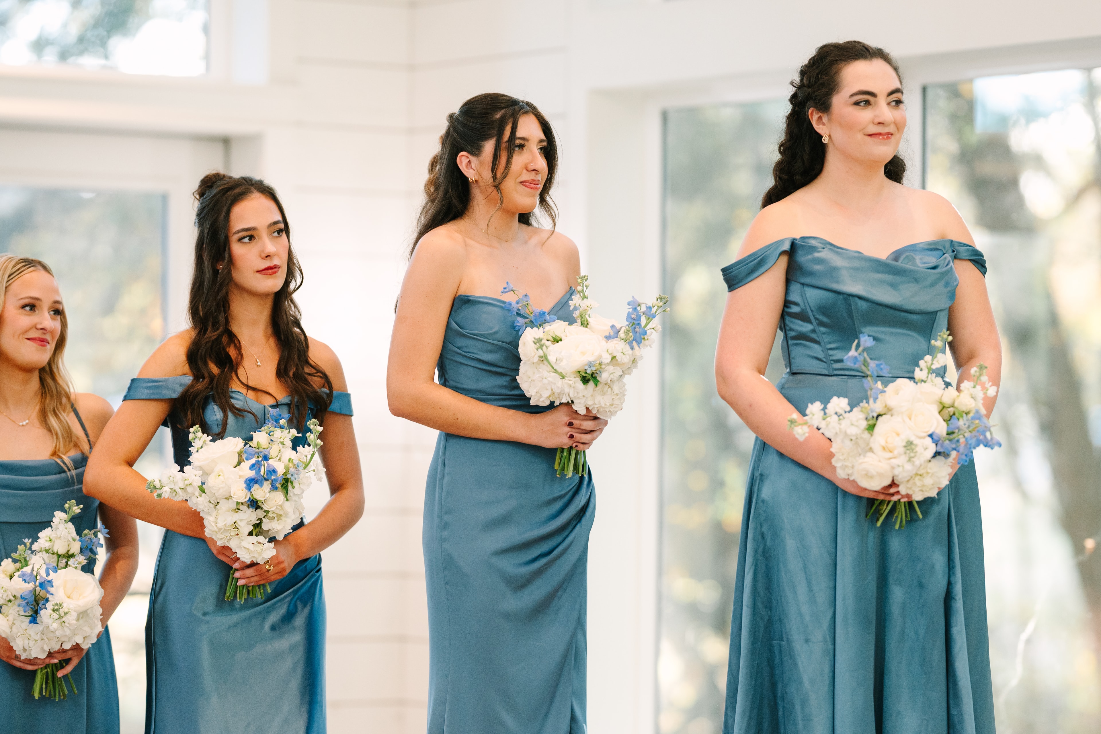 Katie's bridesmaids in steel blue satin off-the-shoulder gowns holding white and blue bouquets, watching her walk down the aisle inside The French Farmhouse chapel