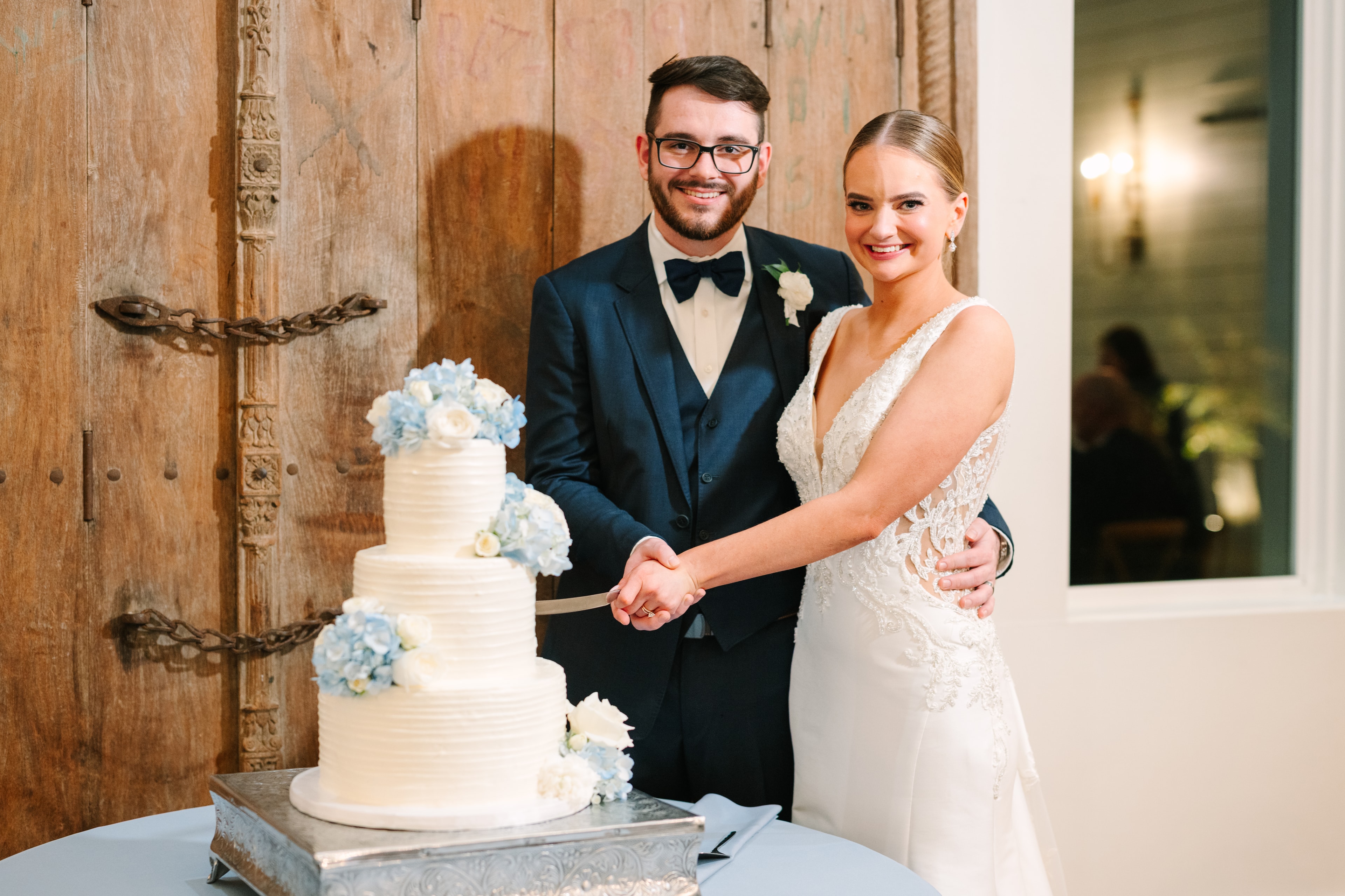 Katie and Mason Allen cutting their three-tier white wedding cake decorated with blue hydrangeas and white roses at The French Farmhouse Venue