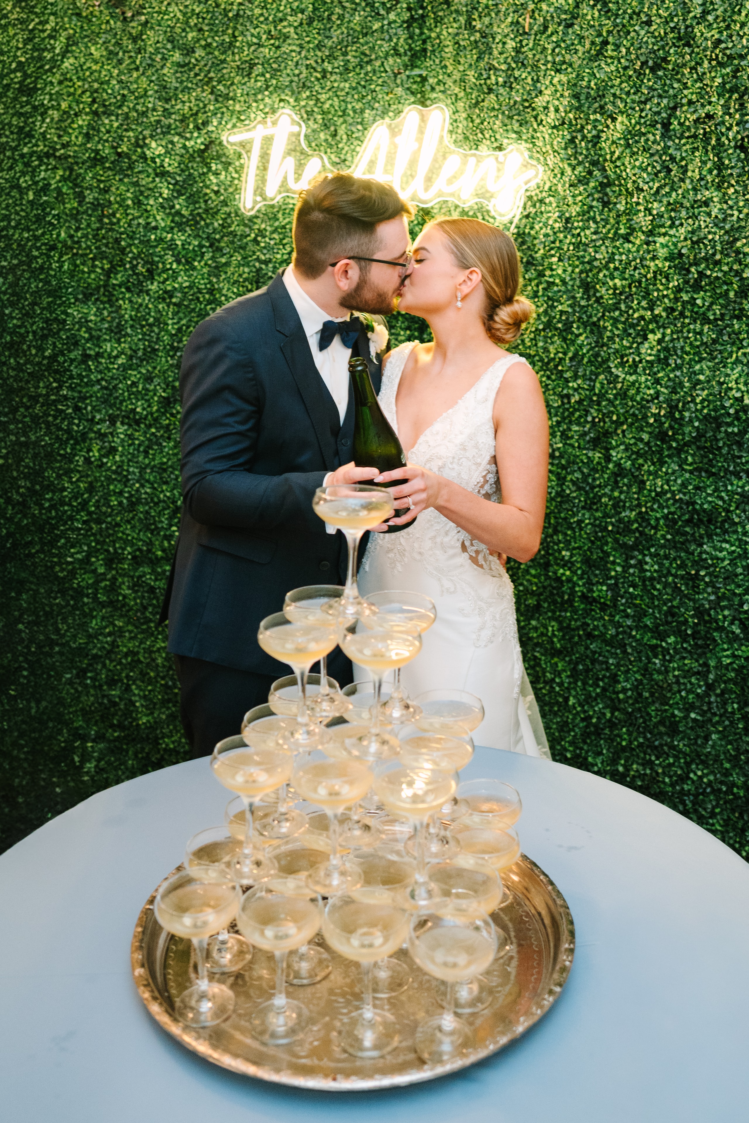 Katie and Mason Allen kissing while pouring champagne down a tower of coupe glasses in front of their glowing 'The Allens' neon sign on a boxwood hedge wall