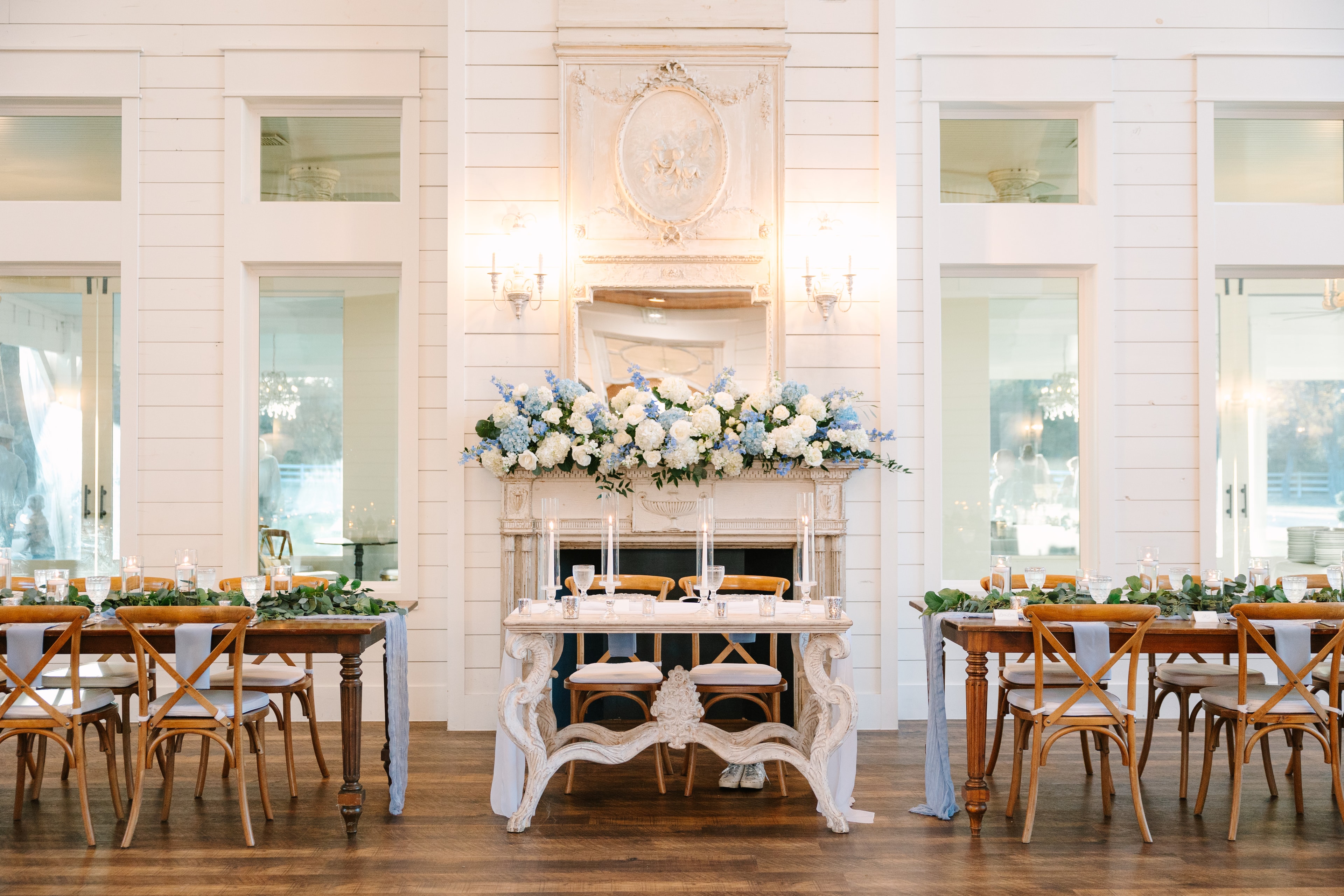 Katie and Mason's reception hall at The French Farmhouse Venue — sweetheart table in front of the ornate white fireplace mantel overflowing with blue and white hydrangeas, farm tables with cross-back chairs and eucalyptus garland runners