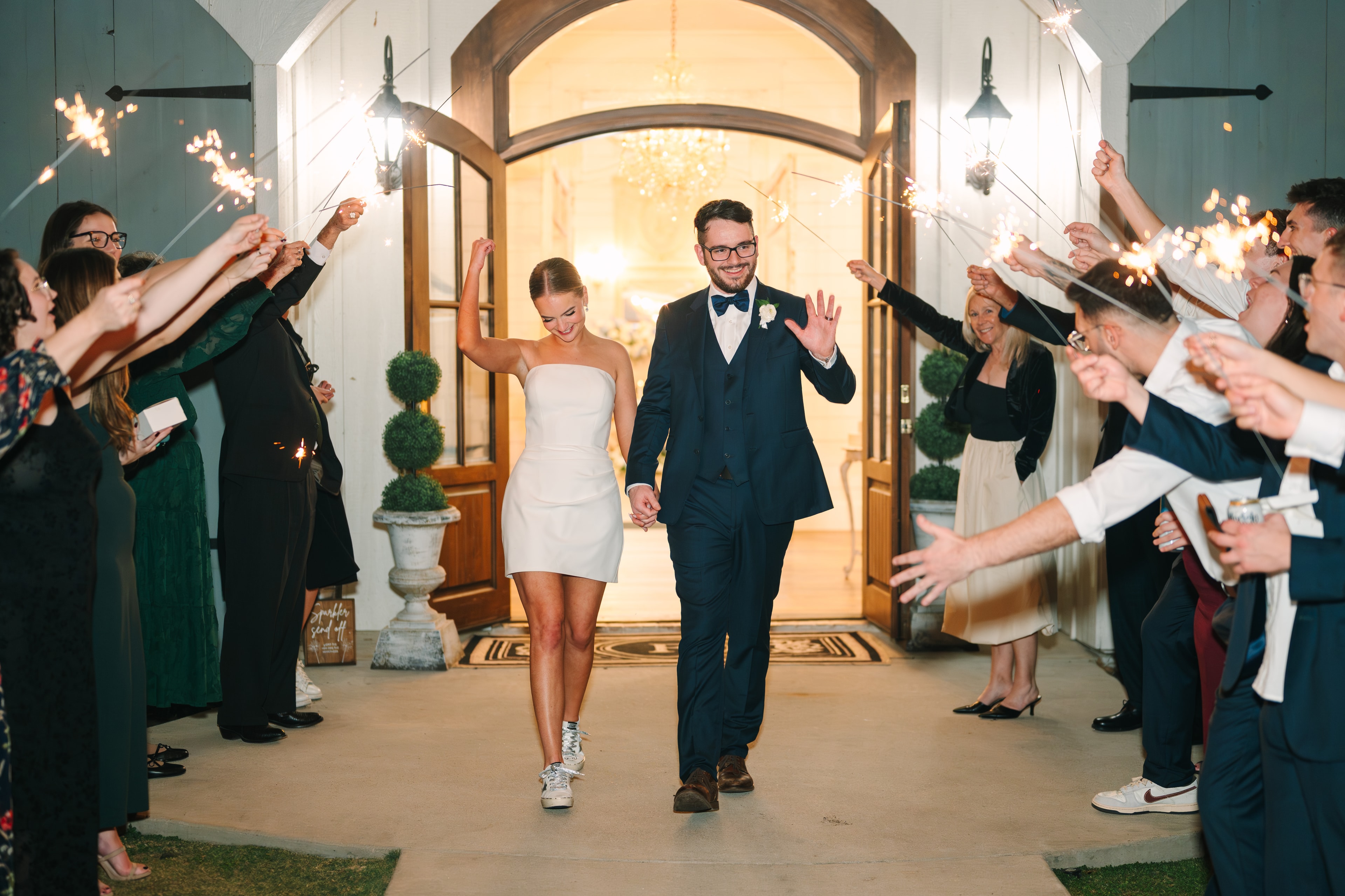 Katie and Mason Allen exiting The French Farmhouse Venue through a sparkler send-off tunnel — Katie in a white mini dress and golden sneakers, Mason in a navy suit, guests lining both sides with sparklers raised under the glowing arched entrance doors