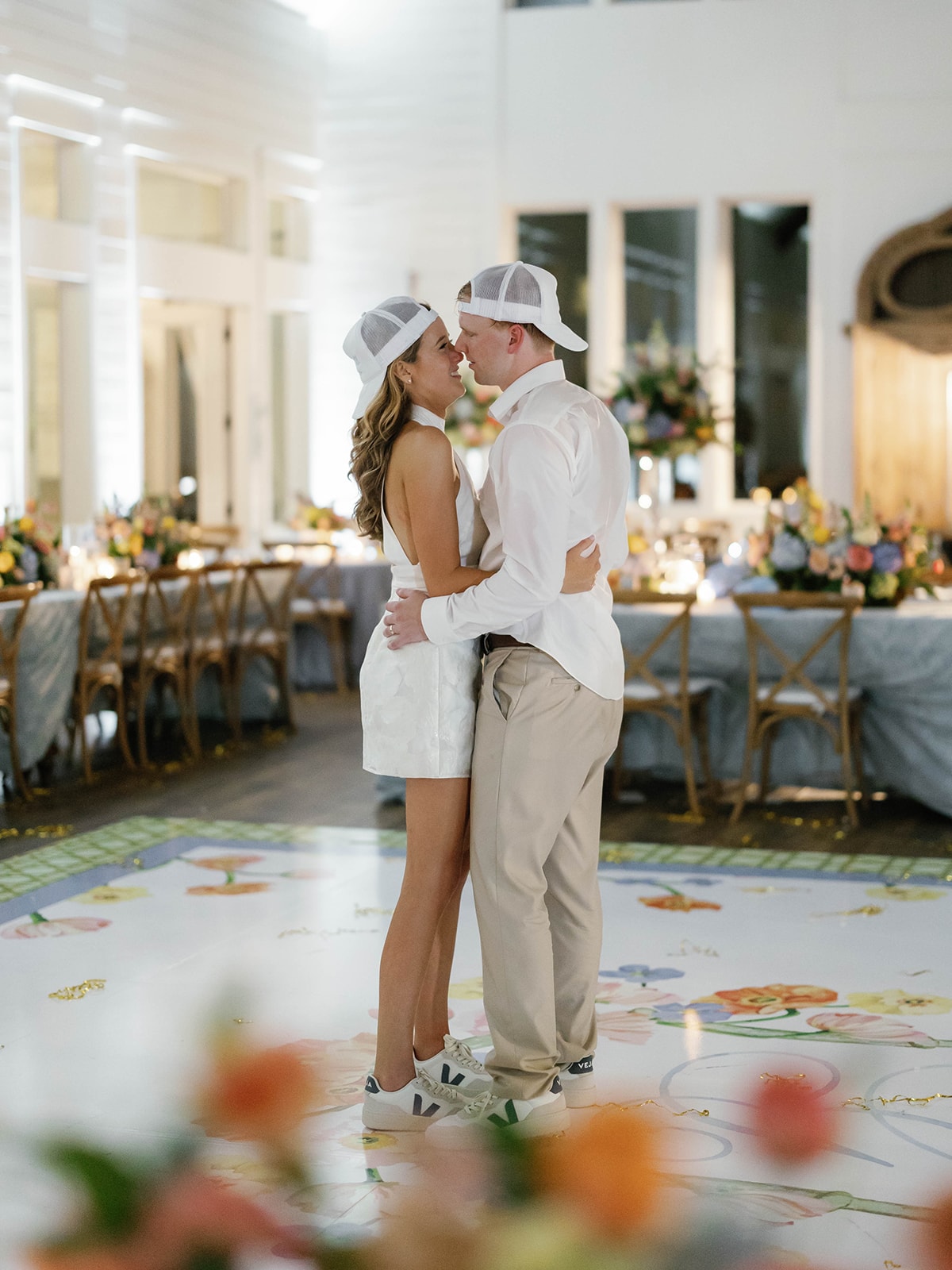 Kylee in a white halter mini dress and Connor in khakis and a white shirt, both wearing white trucker hats, dancing together on the custom floral dance floor