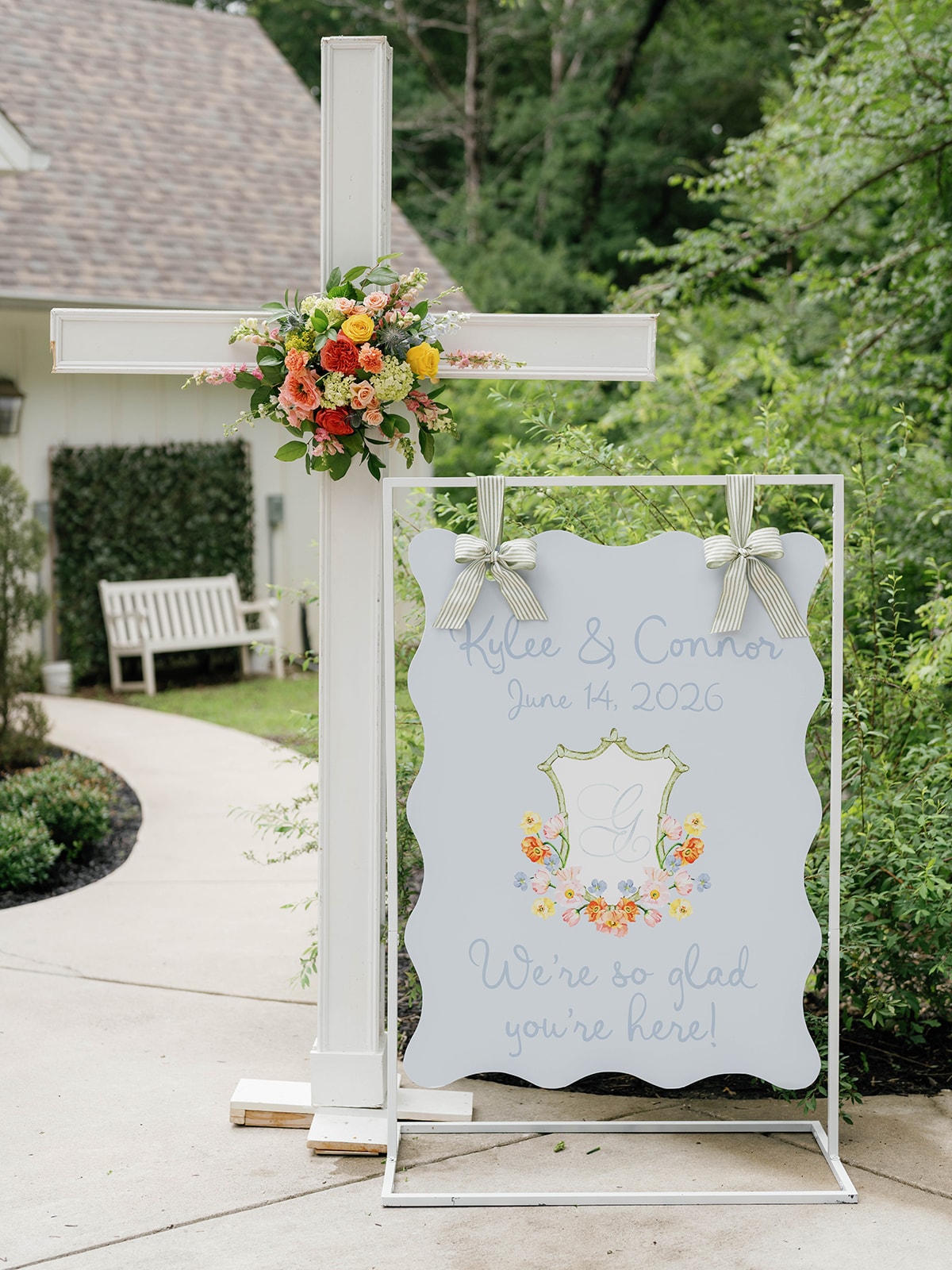 White wooden cross adorned with a colorful wildflower arrangement beside a powder blue scalloped welcome sign reading Kylee and Connor June 14 2026 with striped ribbon bows
