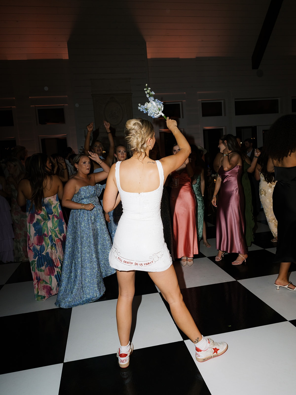 Macey in a short white lace mini dress with spaghetti straps dancing on the dance floor surrounded by guests, arms raised, pure joy on her face