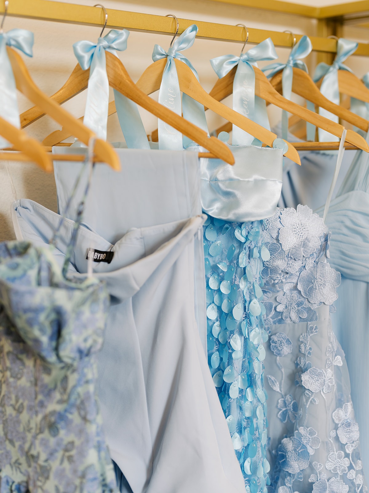 Four dusty rose bridesmaid dresses hanging on a white clothing rack in the bridal suite alongside Macey's white wedding gown