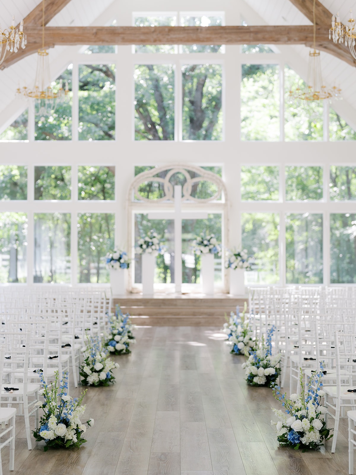 Full view of The French Farmhouse chapel ceremony with guests seated in white Chiavari chairs, lush white floral arrangements lining the aisle, crystal chandeliers overhead, and Macey walking toward Brendan at the ornate white arch