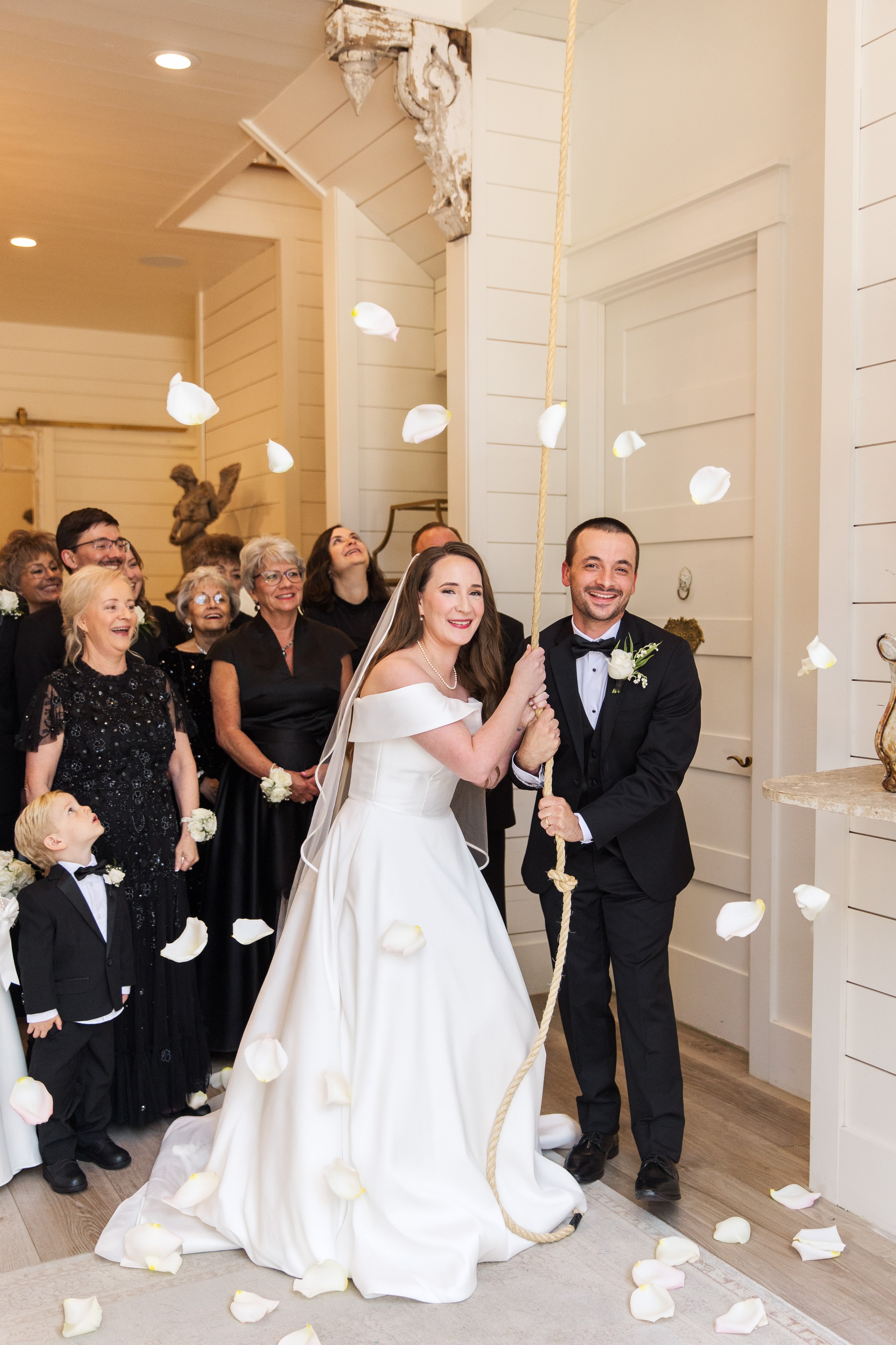 Meaghan and Mason pulling the chapel bell rope together as white rose petals shower down around them, family and the ring bearer watching and laughing in the background, the angel statue and white shiplap walls of The French Farmhouse behind them