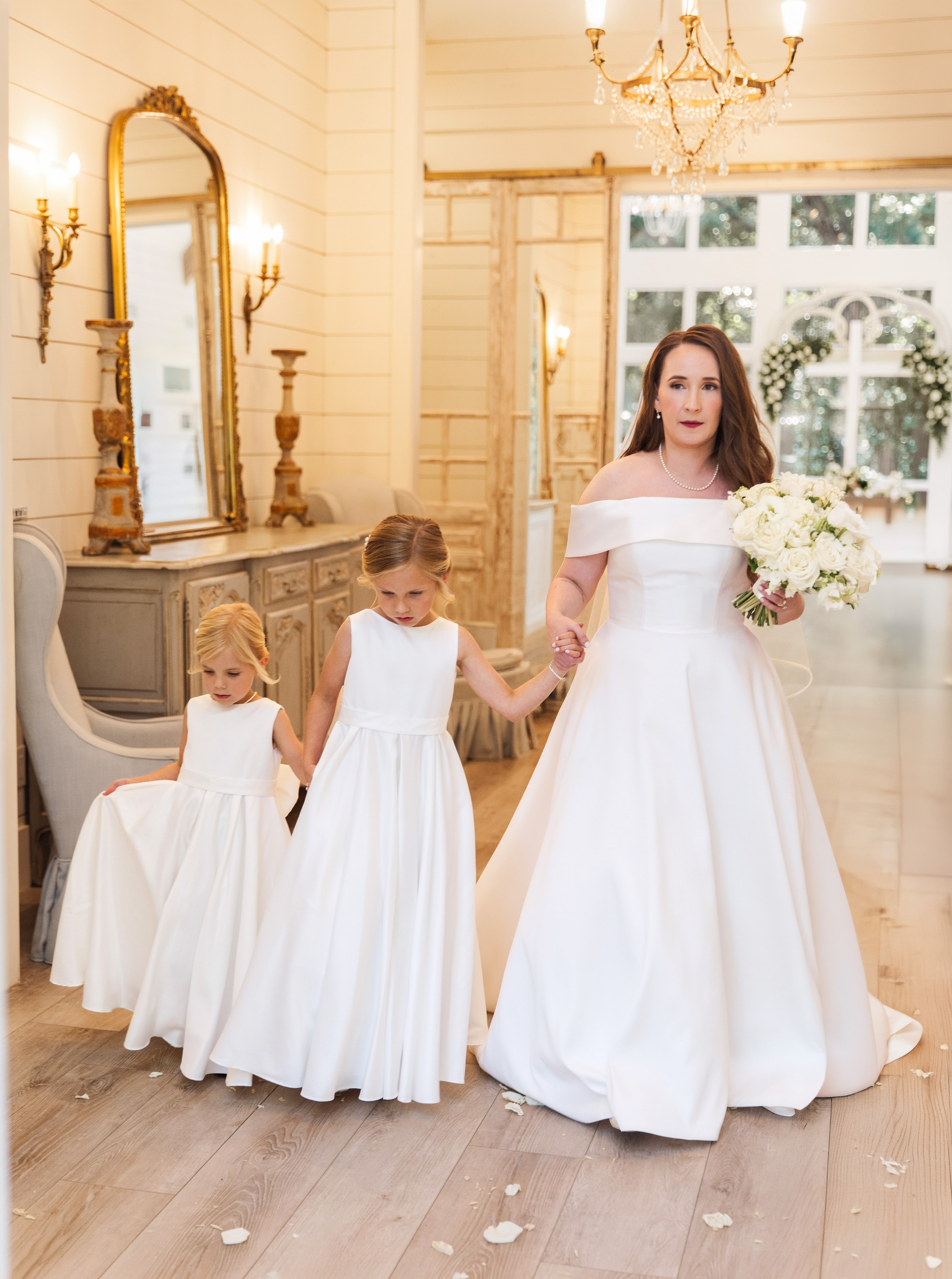 Meaghan in her off-the-shoulder white ballgown holding hands with two flower girls in matching white dresses inside the French Farmhouse bridal suite, the gold ornate mirror, crystal chandelier, and white floral arch visible behind them