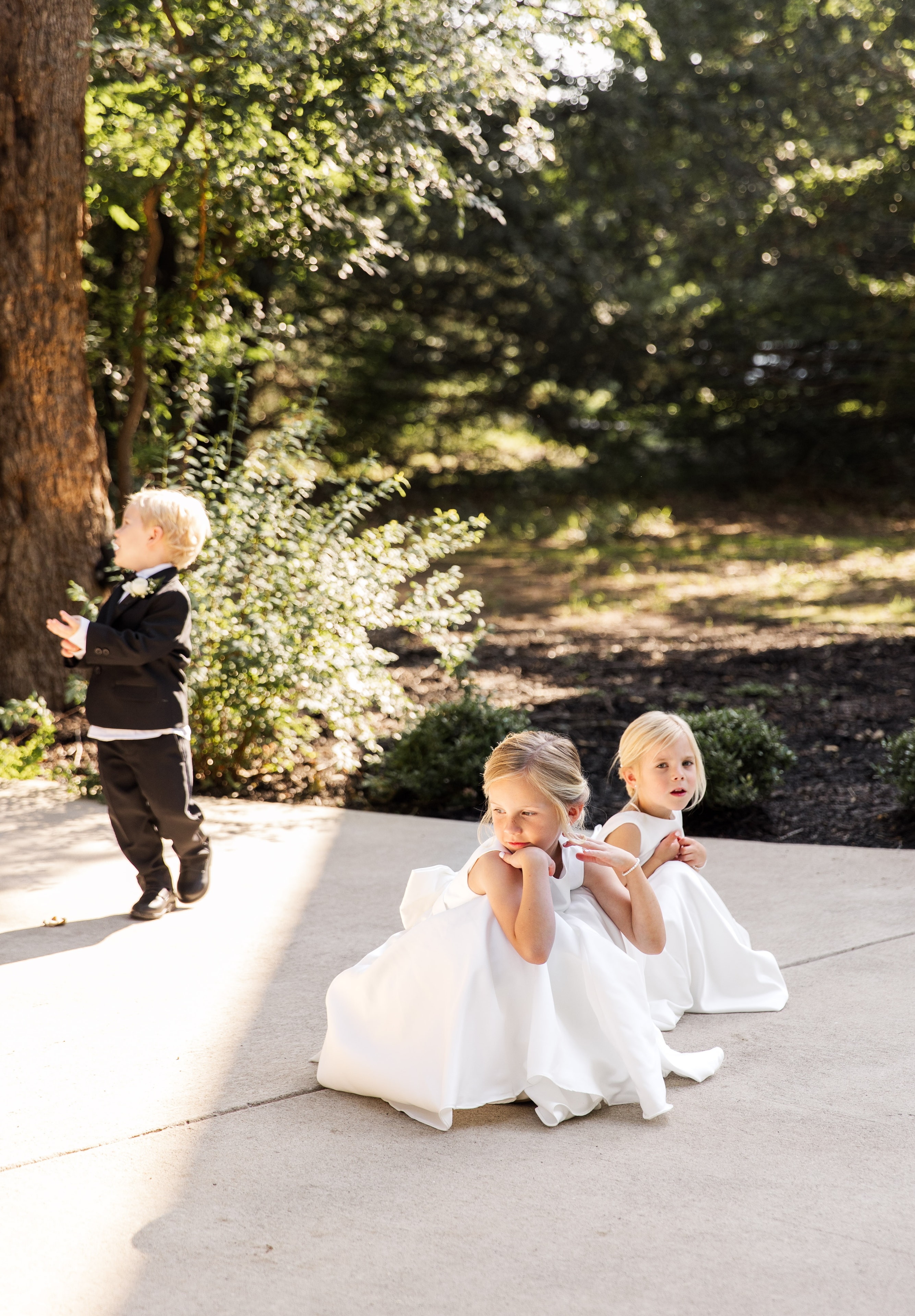 Two flower girls in white dresses sitting on the sunny courtyard pavement looking bored and adorable, a little ring bearer in a black tuxedo playing behind them against a tree on the French Farmhouse grounds