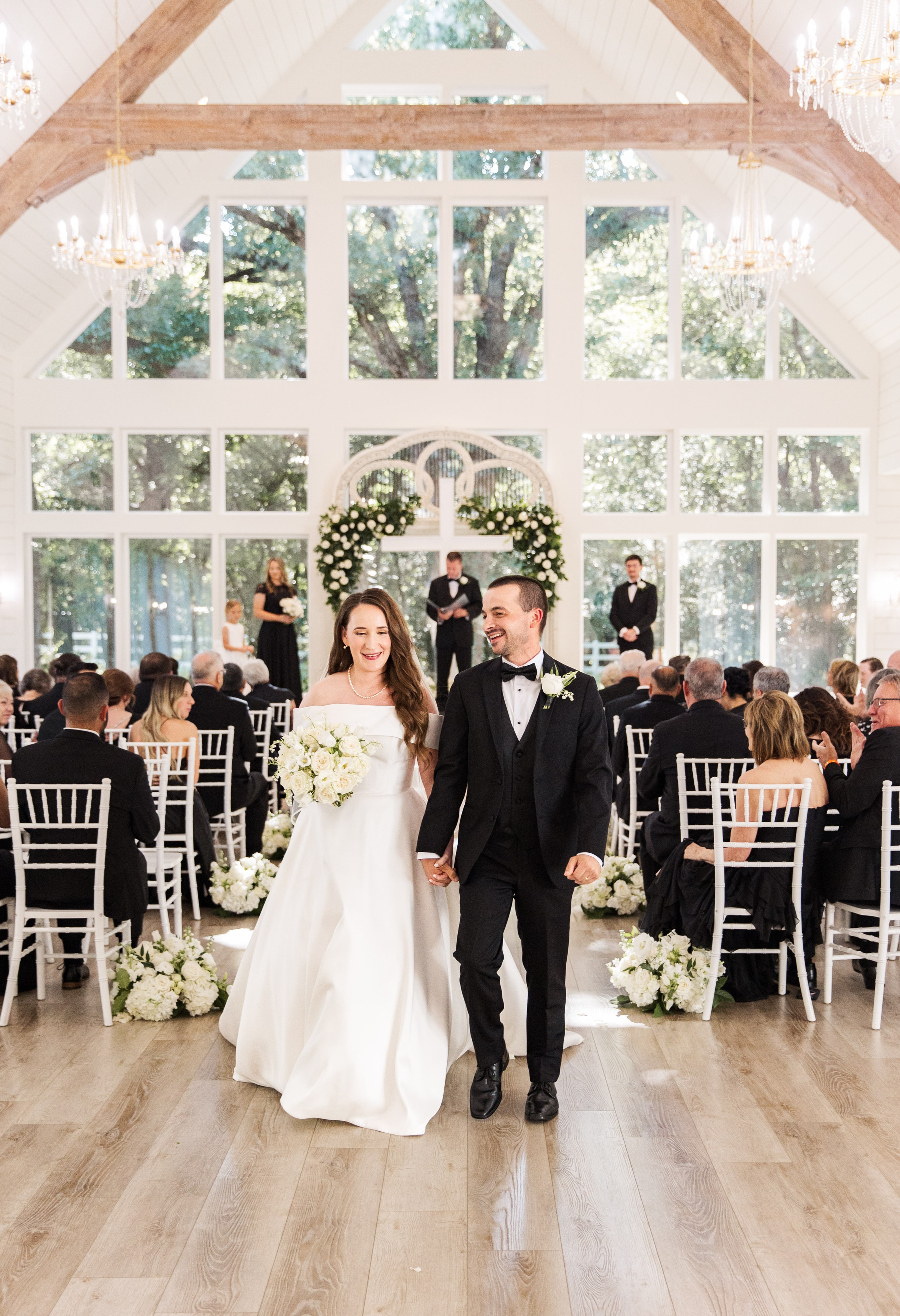 Meaghan and Mason beaming as they walk back down the aisle together as newlyweds inside The French Farmhouse chapel, white hydrangea and rose arrangements lining the aisle, guests cheering on both sides, the antique arch and floor-to-ceiling windows glowing behind them