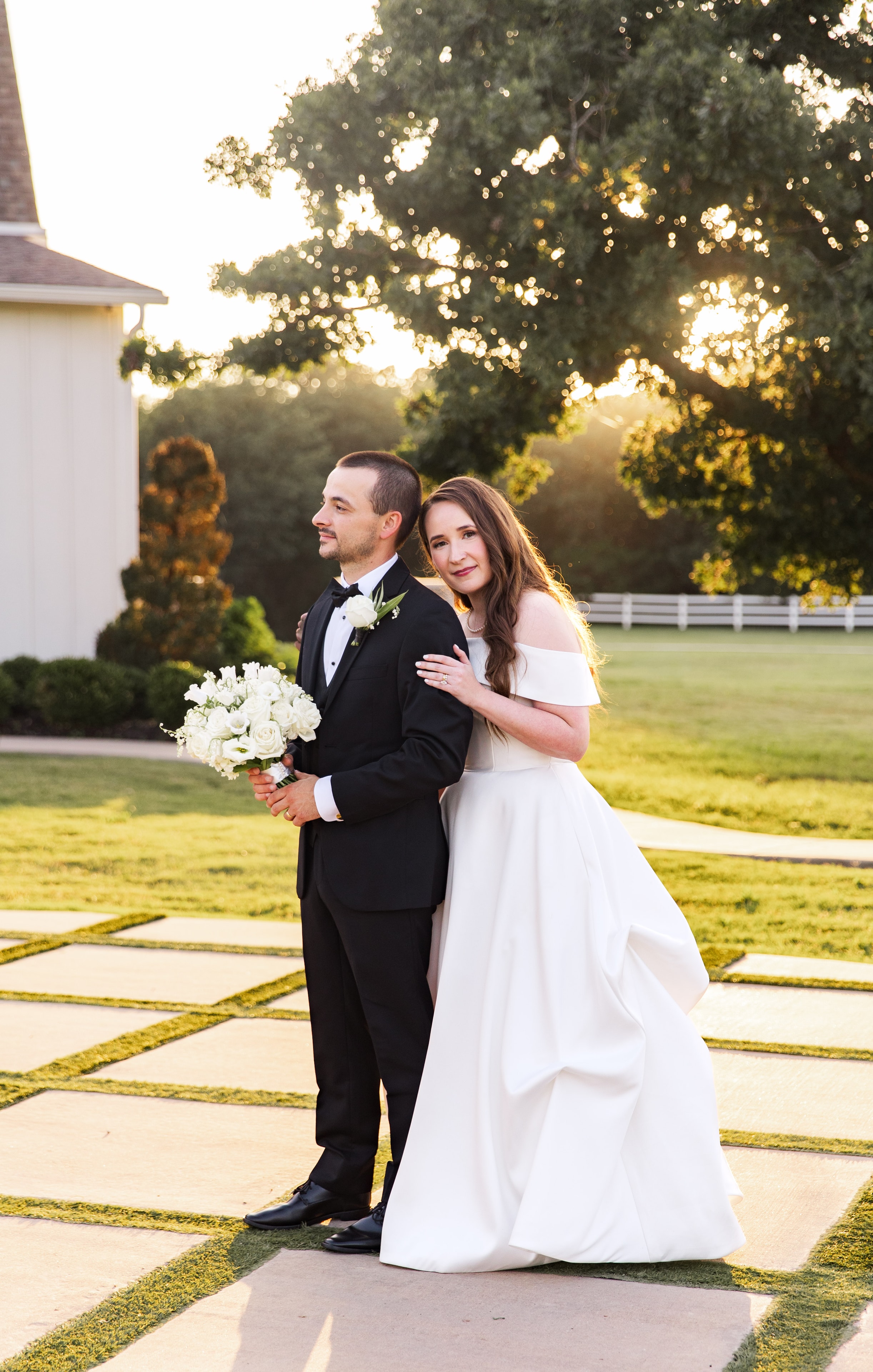 Meaghan leaning against Mason's back on the checkerboard courtyard at golden hour, both looking away from each other with soft smiles, Meaghan holding her white rose bouquet, the white pavilion and white fence glowing in the warm evening light behind them