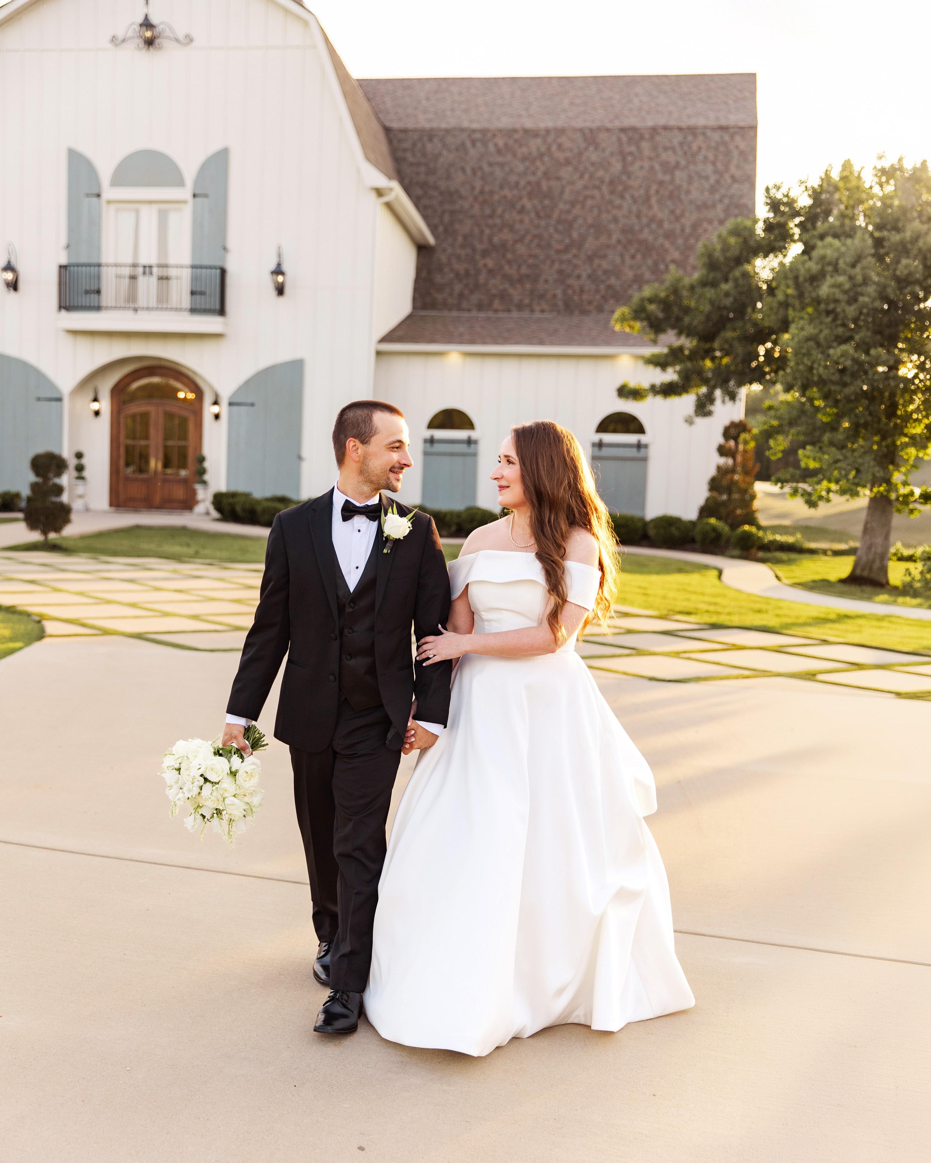 Meaghan and Mason walking hand in hand across the checkerboard courtyard in front of the white French Farmhouse barn at golden hour, both laughing and looking at each other, the sage shutters and arched wood doors glowing behind them