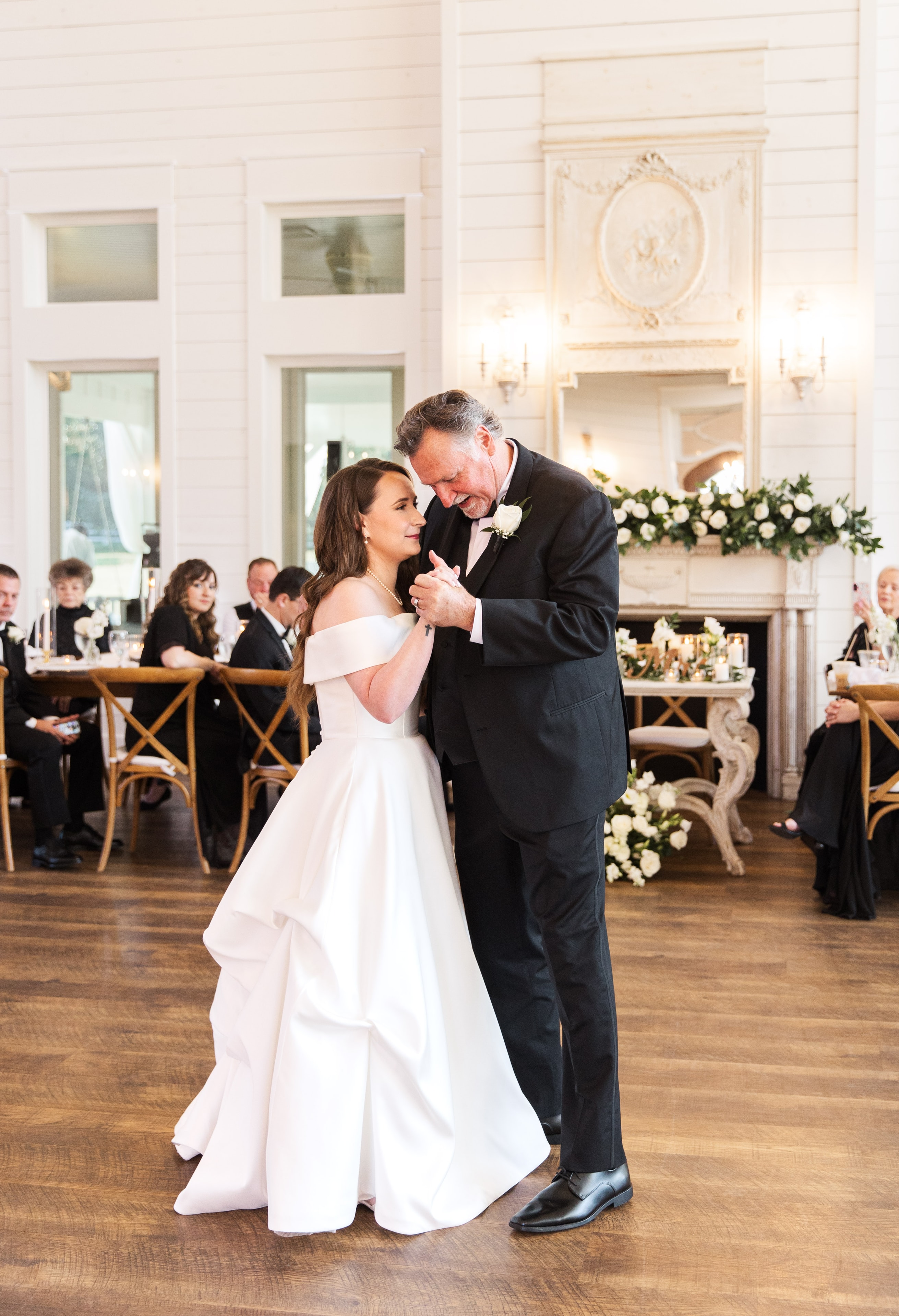 Meaghan sharing a tender father-daughter dance inside The French Farmhouse reception hall, her off-the-shoulder white ballgown sweeping the hardwood floor, the ornate white fireplace mantle with lush white rose and greenery garland glowing behind them, guests seated at cross-back chairs watching on both sides