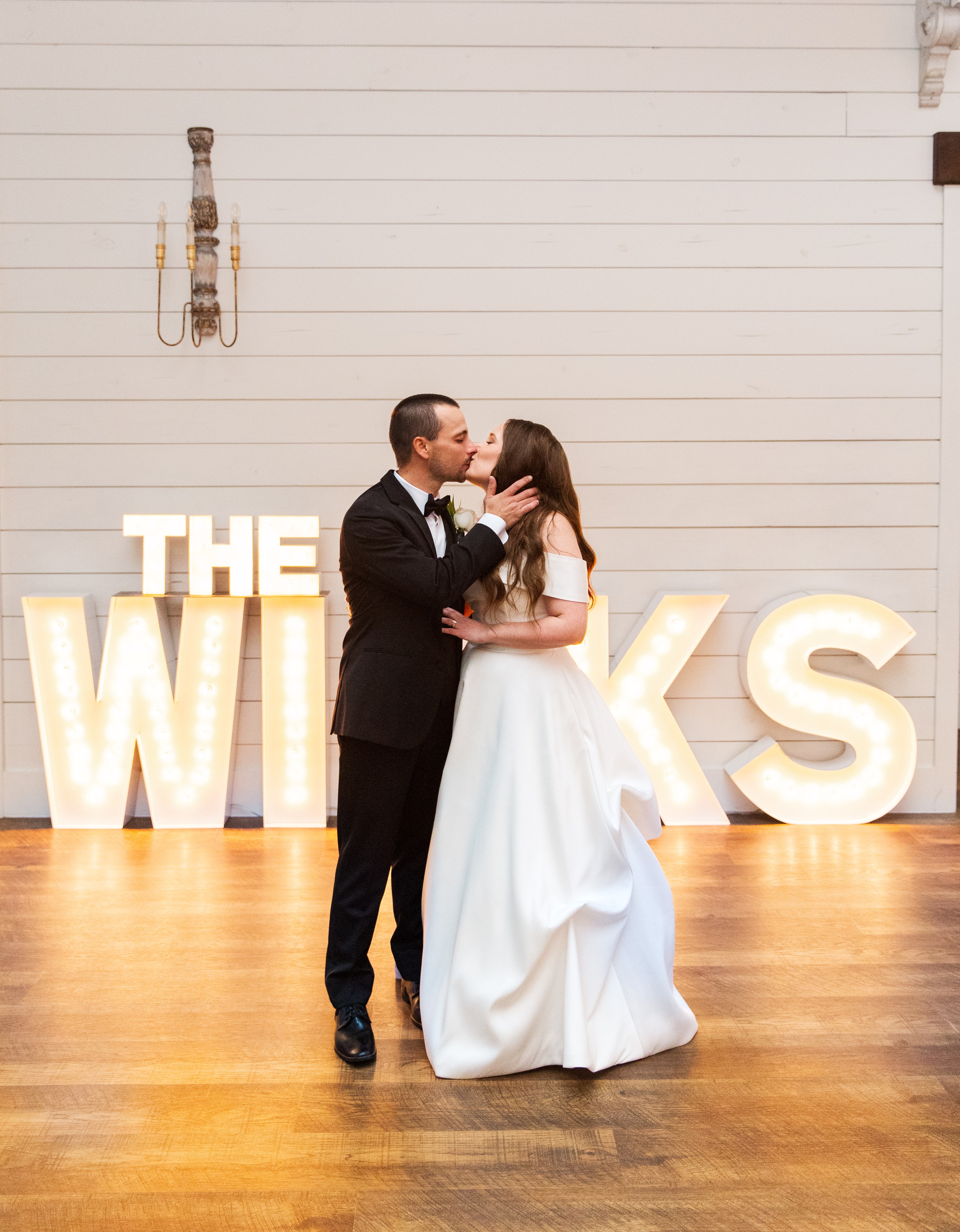 Meaghan and Mason sharing a kiss on the dance floor in front of the glowing THE WILKS marquee letter lights against the white shiplap wall of The French Farmhouse reception hall, the warm glow of the marquee letters reflecting on the hardwood floor around them