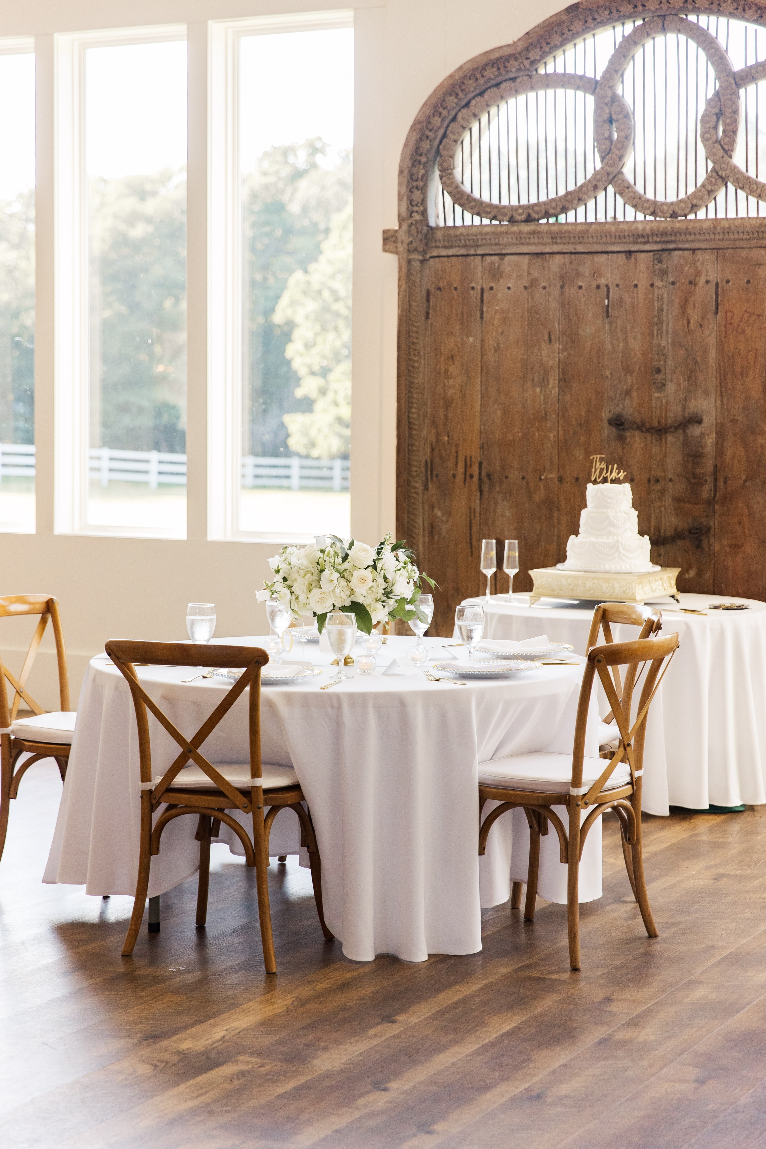 Elegant reception tablescape inside The French Farmhouse reception hall — white linen round table with white rose and greenery centerpiece, cross-back wooden chairs, gold flatware, and The Wilks wedding cake displayed on a white table in front of the antique carved wood barn doors, floor-to-ceiling windows overlooking the white fence and green grounds beyond