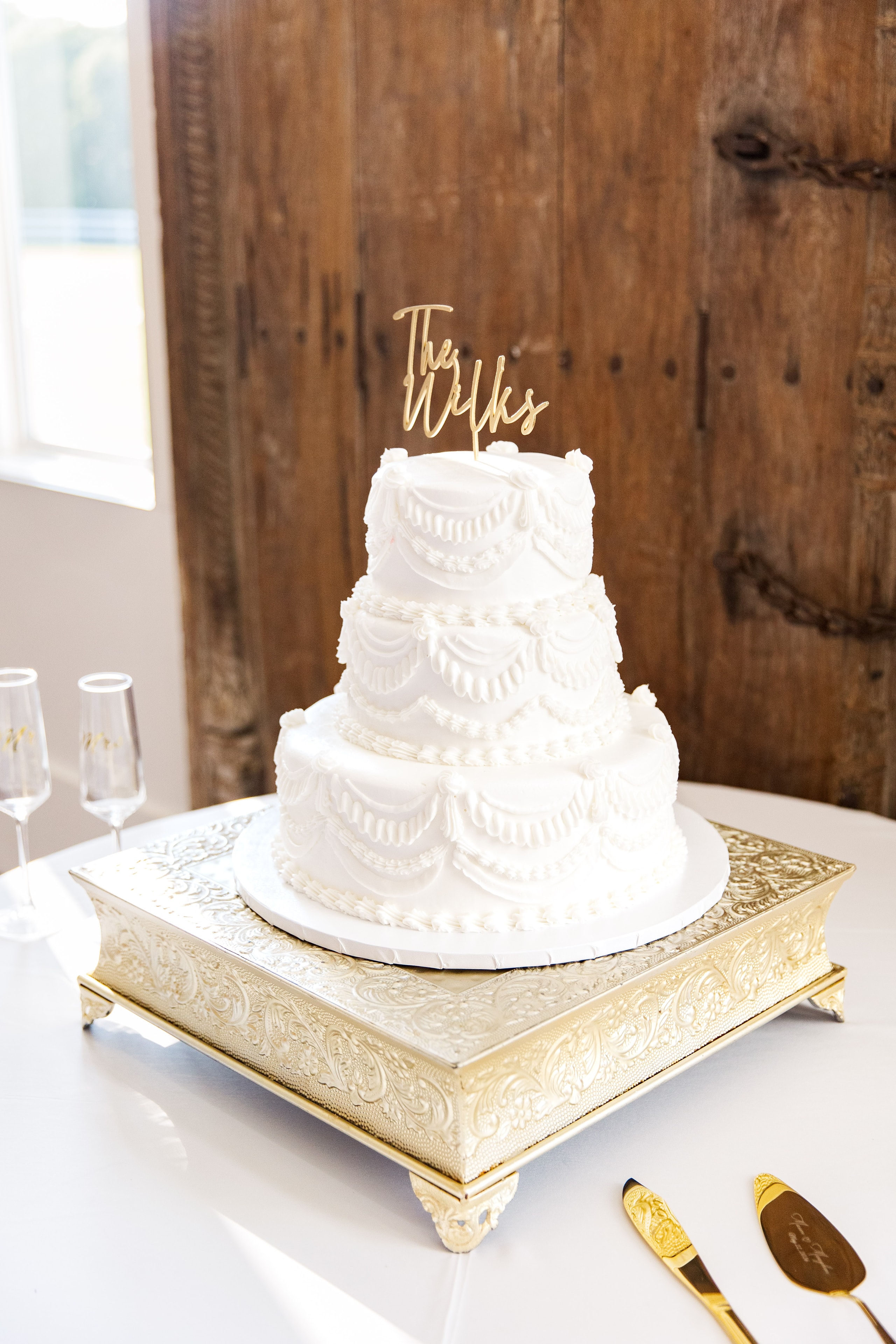 Three-tier white textured wedding cake with intricate piped draping and floral details, topped with a gold script The Wilks cake topper, displayed on an ornate gold square cake stand with gold cake cutting set beside it, the antique carved wood barn doors of The French Farmhouse reception hall behind it