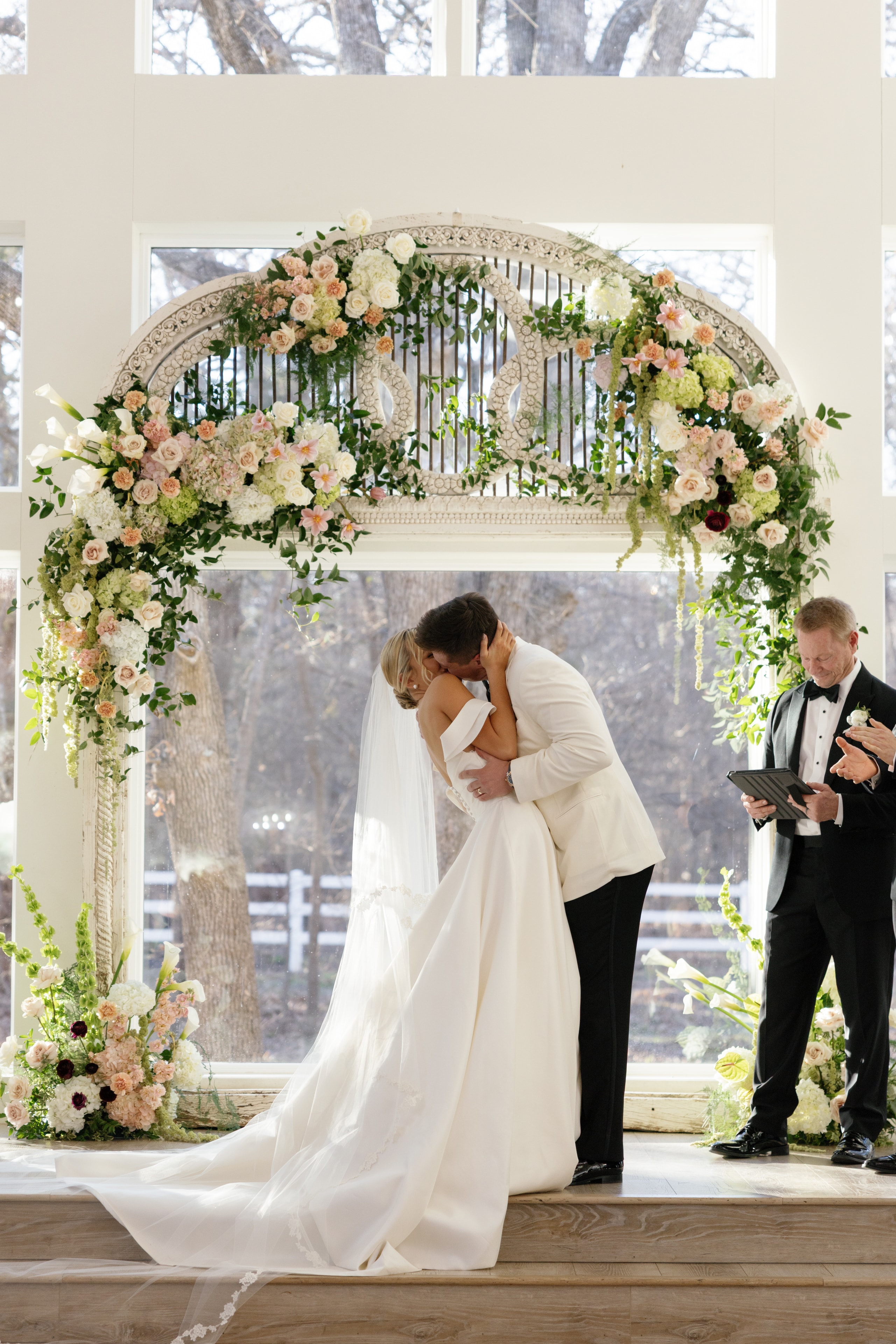 Megan and Rex sharing their first kiss under the lush floral antique arch at The French Farmhouse chapel