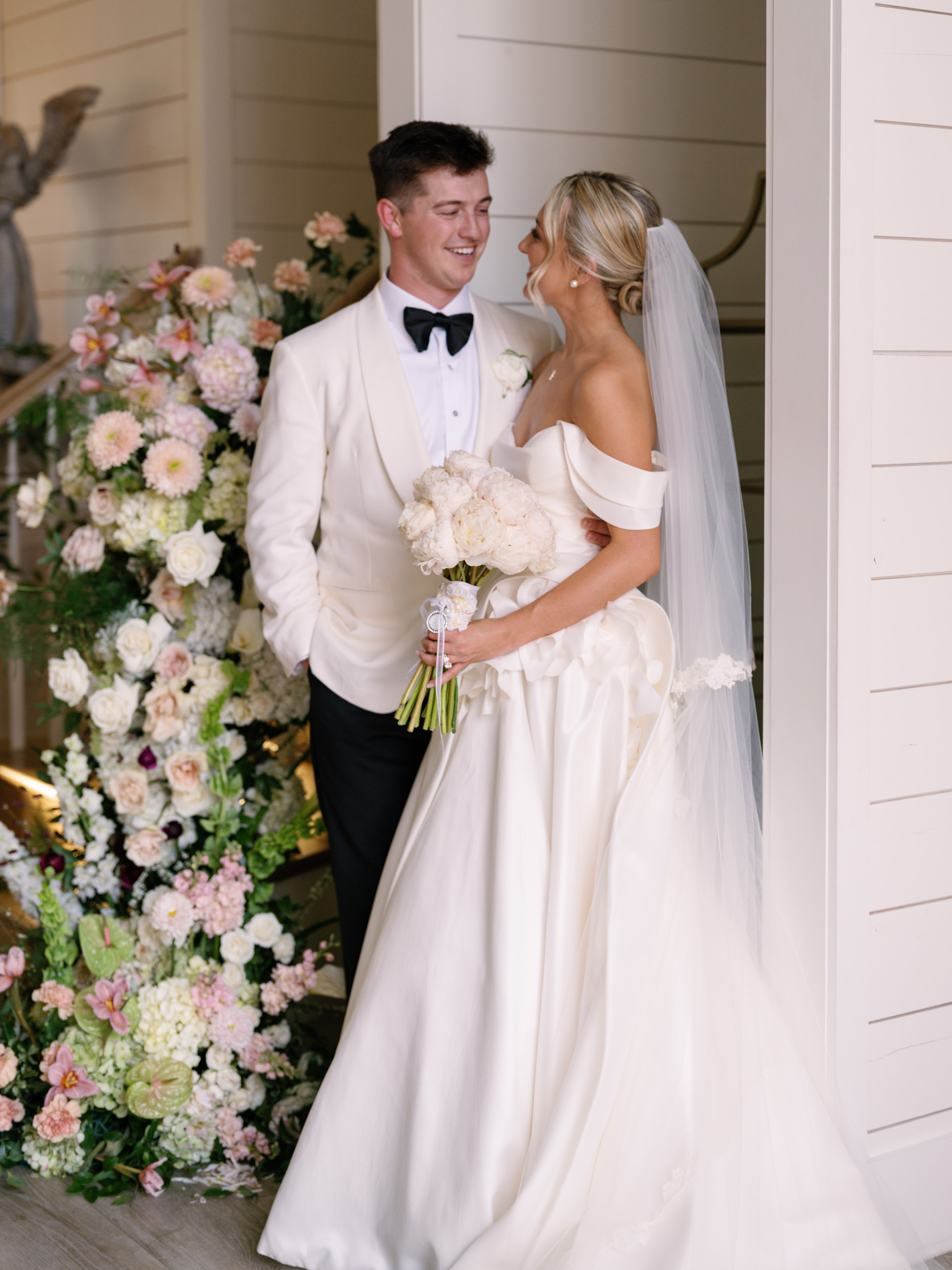 Megan and Rex laughing together by the lush pink and white floral staircase installation at The French Farmhouse Venue