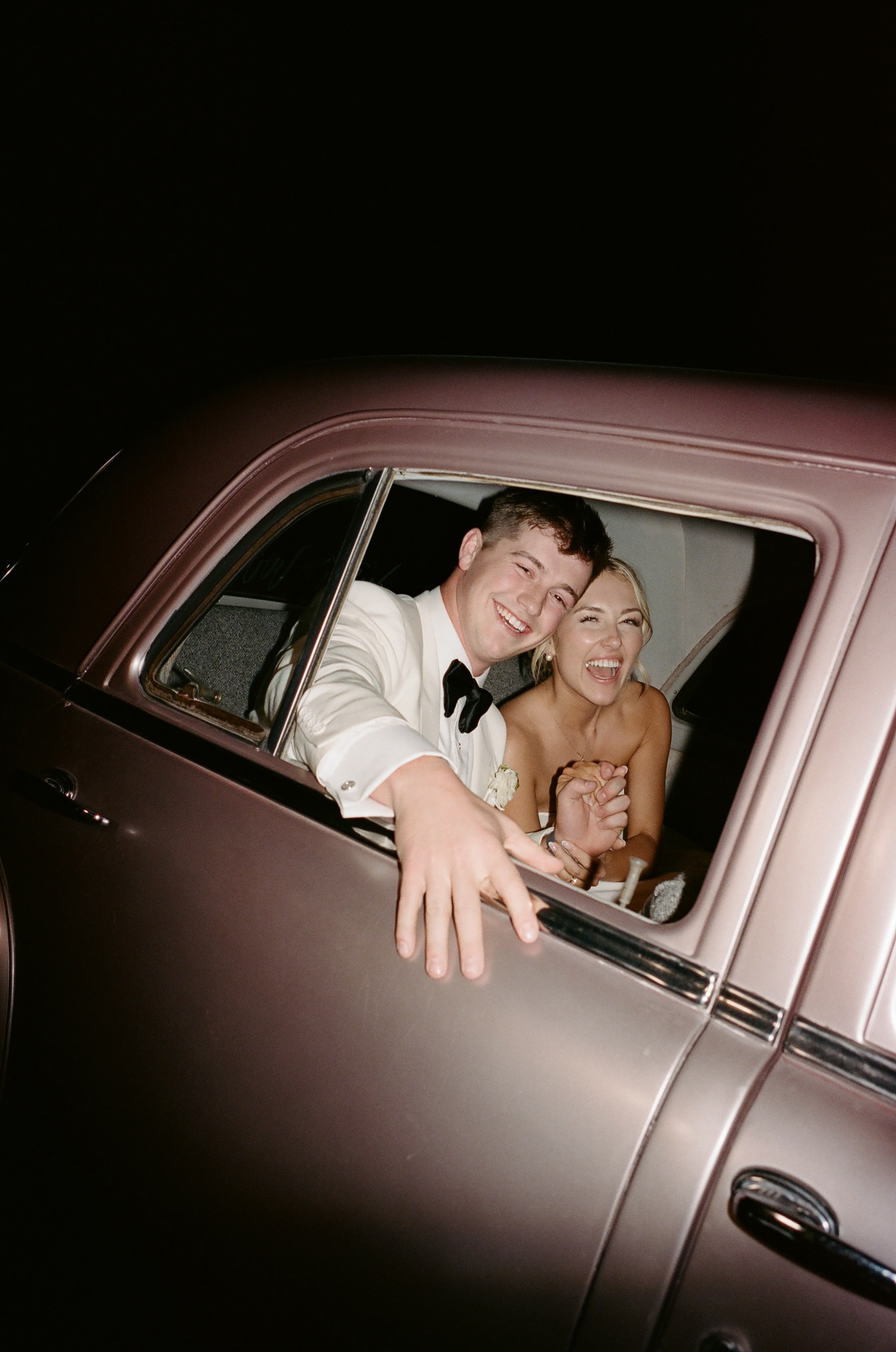 Megan and Rex laughing and waving from the window of their vintage getaway car