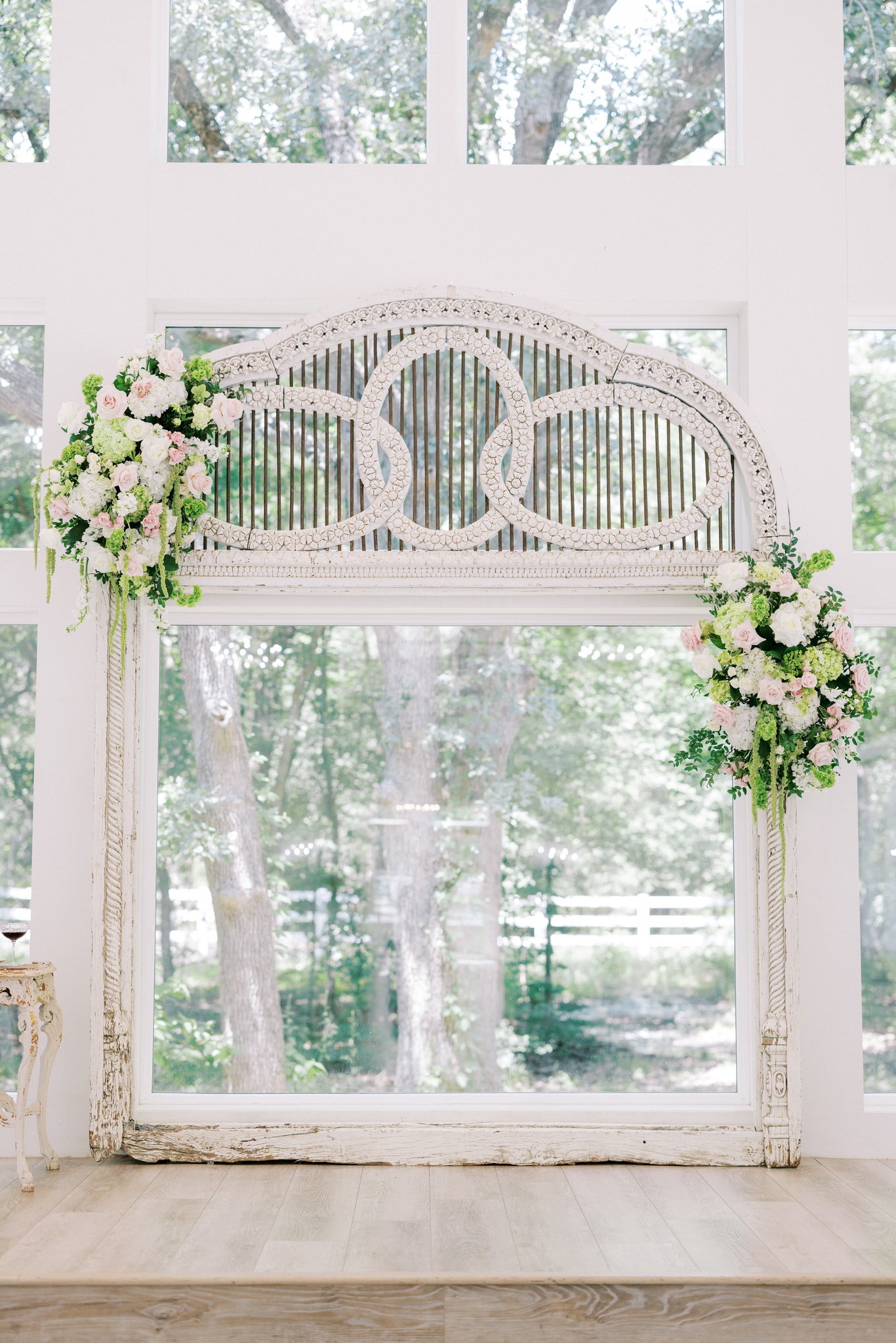 The French Farmhouse antique arch adorned with blush pink roses, white hydrangea, and trailing green amaranthus on both sides, set against the floor-to-ceiling chapel windows with mature oak trees beyond