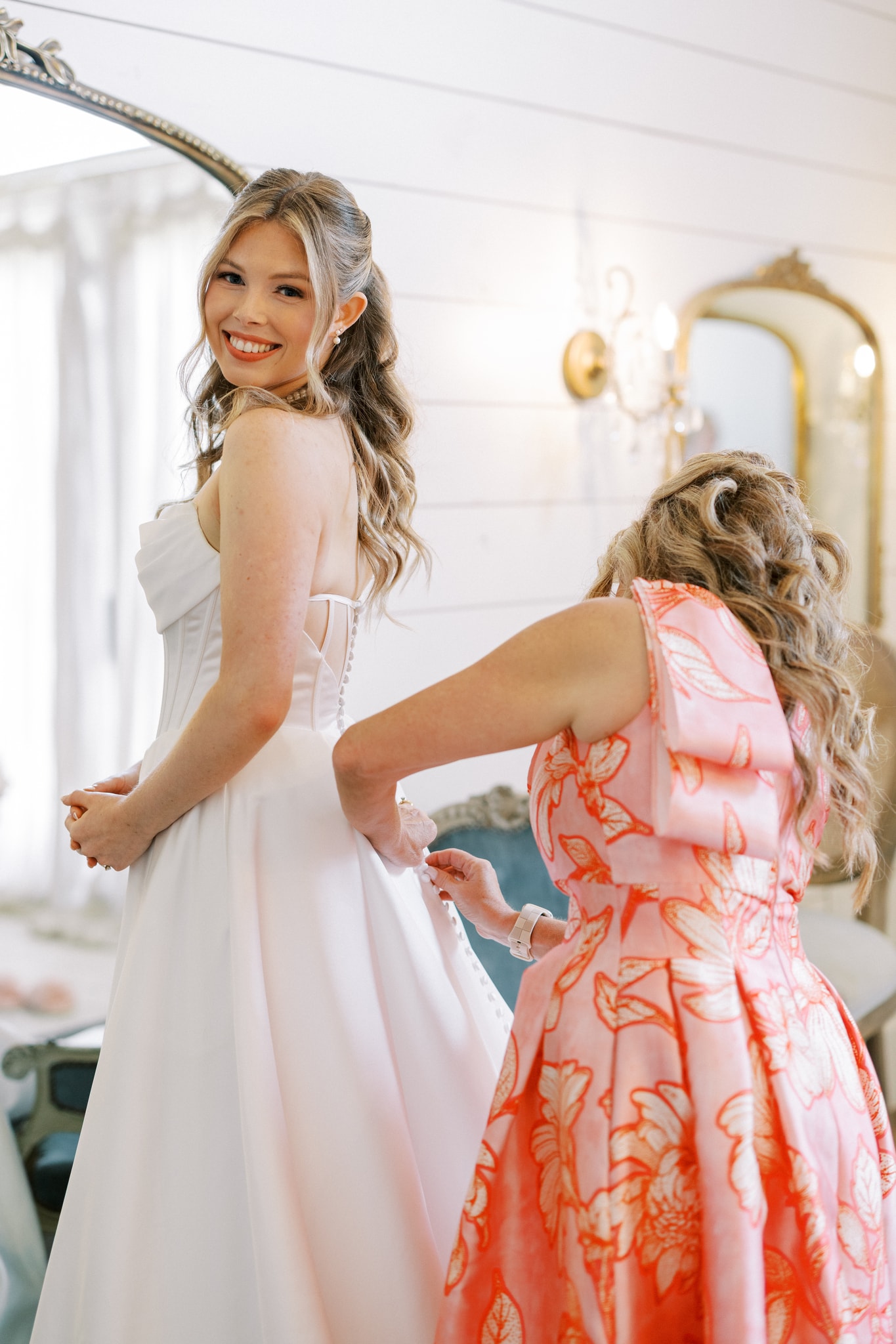 Morgan smiling over her shoulder in the bridal suite as someone buttons the back of her elegant white gown, the ornate gold mirror and sconce lighting behind her
