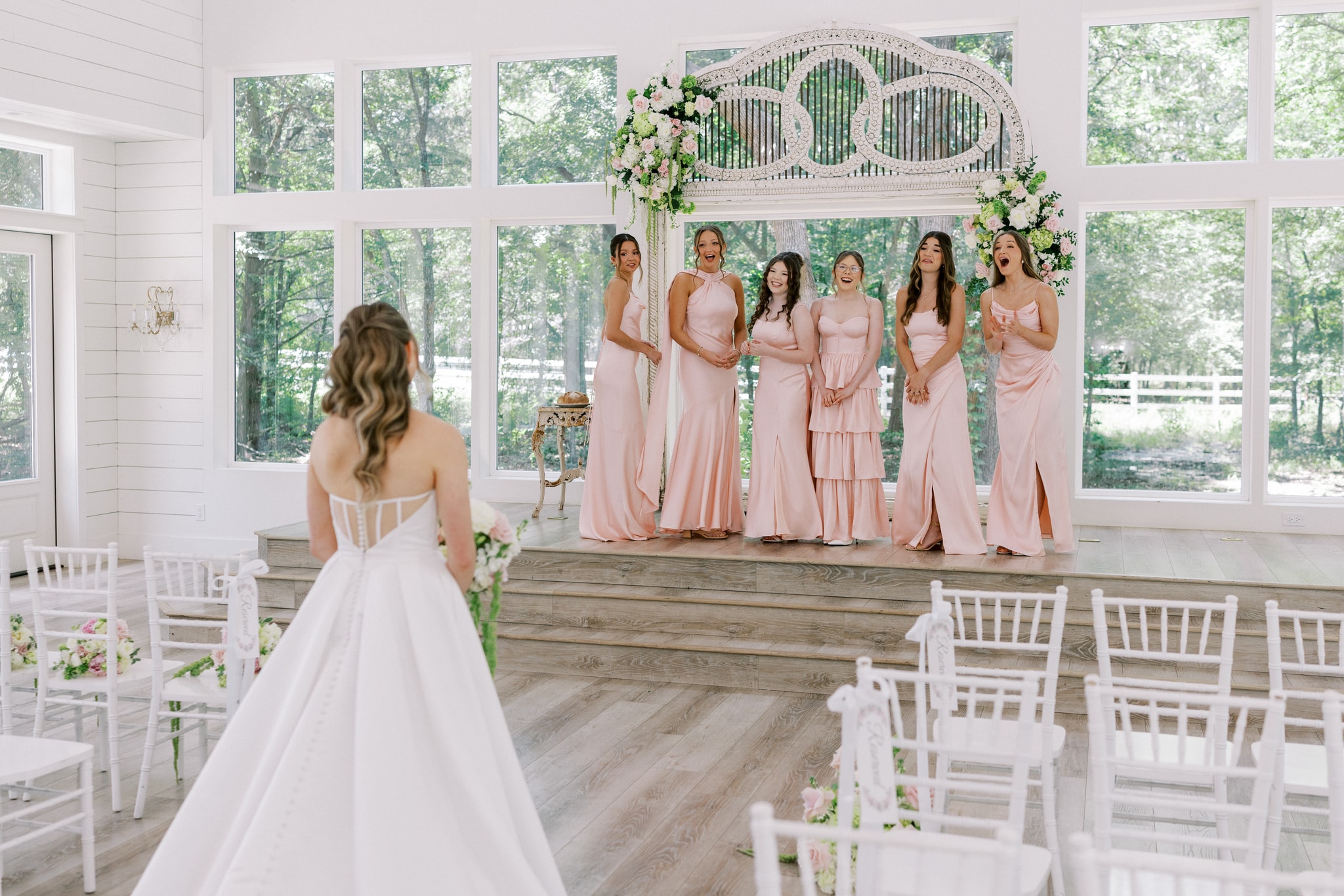 Morgan in her full bridal gown walking toward her bridesmaids in blush dresses standing on the chapel stage — all six bridesmaids gasping and screaming with joy at their first look