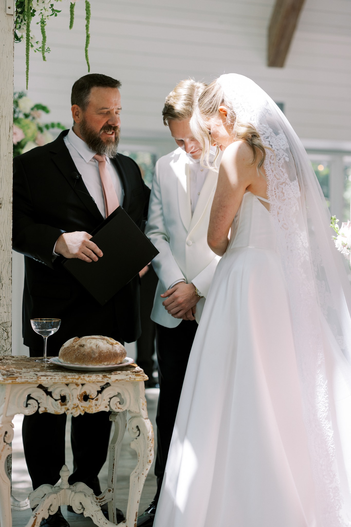Morgan bowing her head during the ceremony as the officiant reads from his book beside her and Jack, a rustic white ornate table holding a loaf of bread and a crystal wine glass in the foreground