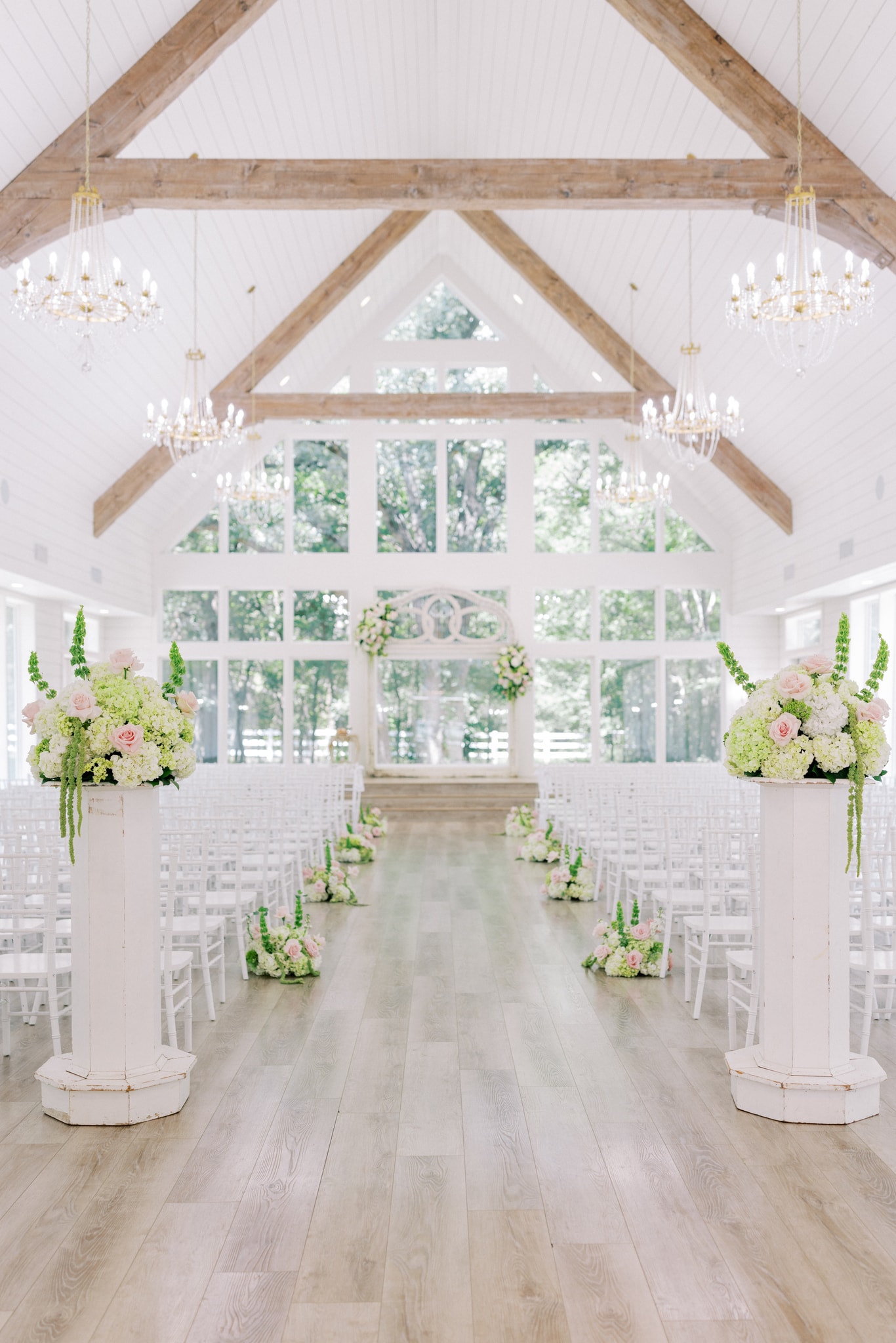 The French Farmhouse chapel set for Morgan and Jack's ceremony — white Chiavari chairs, tall pedestal arrangements of blush roses and white hydrangea, exposed wood beam trusses, crystal chandeliers, and floor-to-ceiling windows with trees beyond