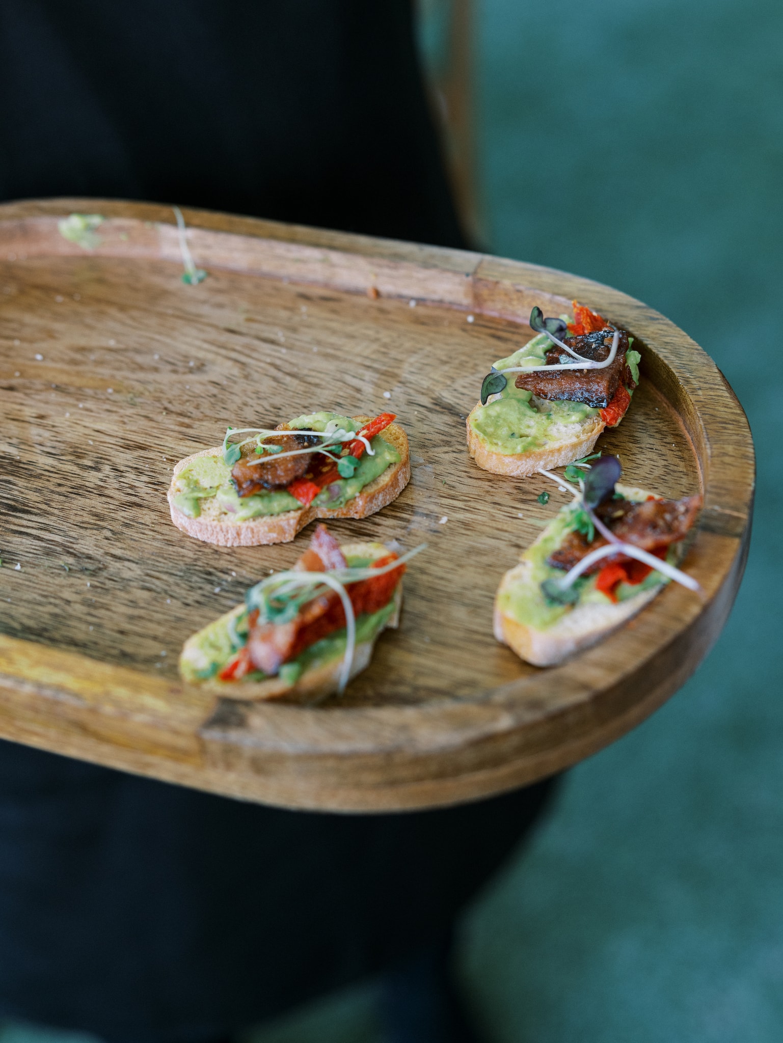 A server holding a round wooden board with avocado bruschetta bites topped with roasted tomatoes and microgreens during cocktail hour