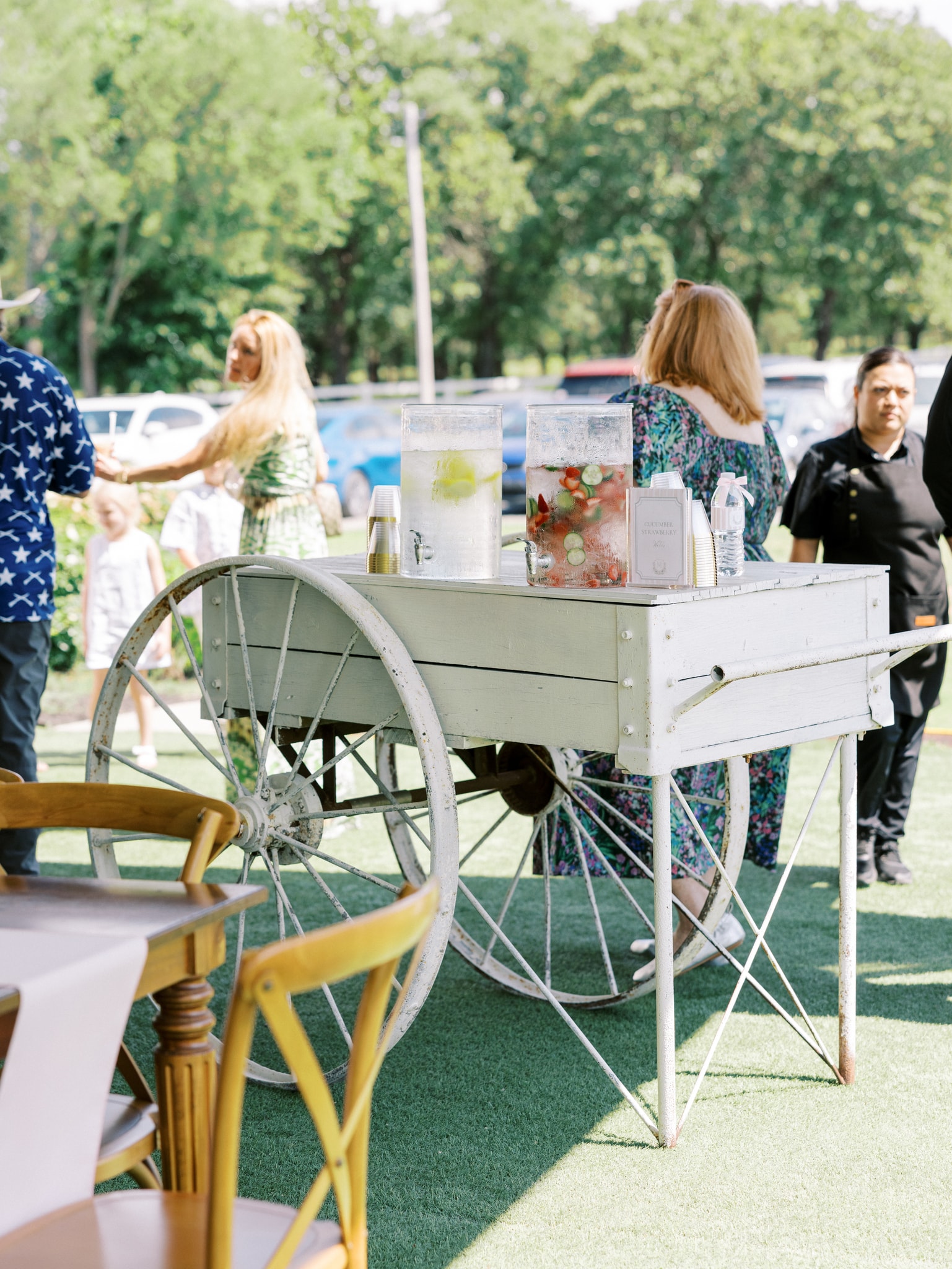 A vintage white wooden drink cart with large glass dispensers of infused water and lemonade on the lawn during cocktail hour, guests mingling in the background under mature oak trees