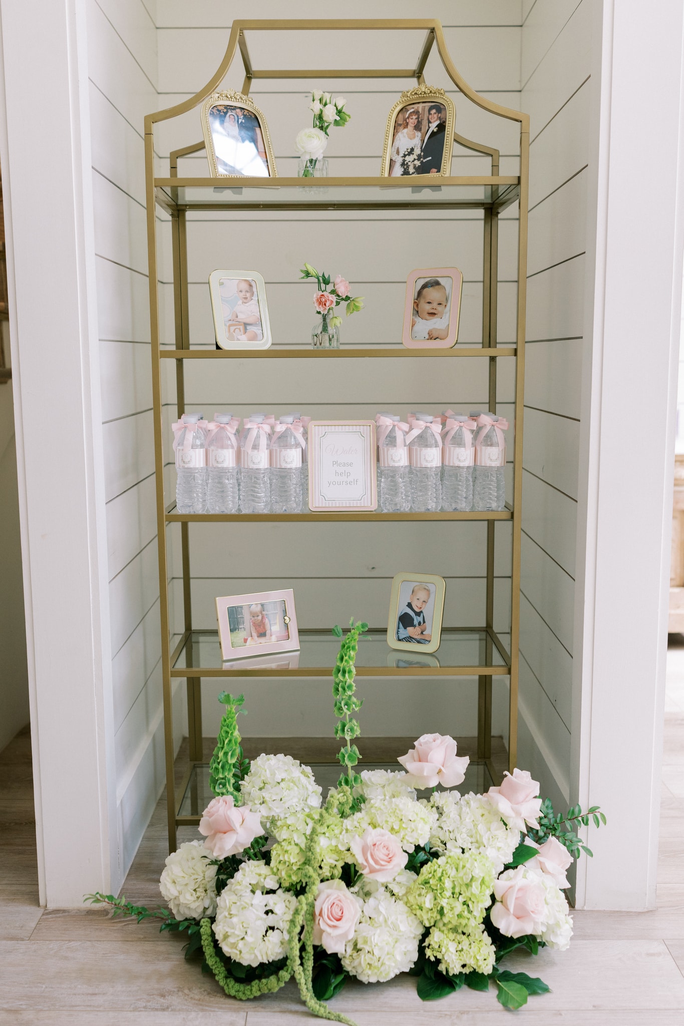 A gold etagere display shelf in the French Farmhouse venue hallway — parents' wedding photos in ornate frames on top, baby photos of Morgan and Jack in pink and green frames on the shelves, custom water bottles with pink bows, and a lush arrangement of blush roses, white hydrangea, and green foxglove at the base
