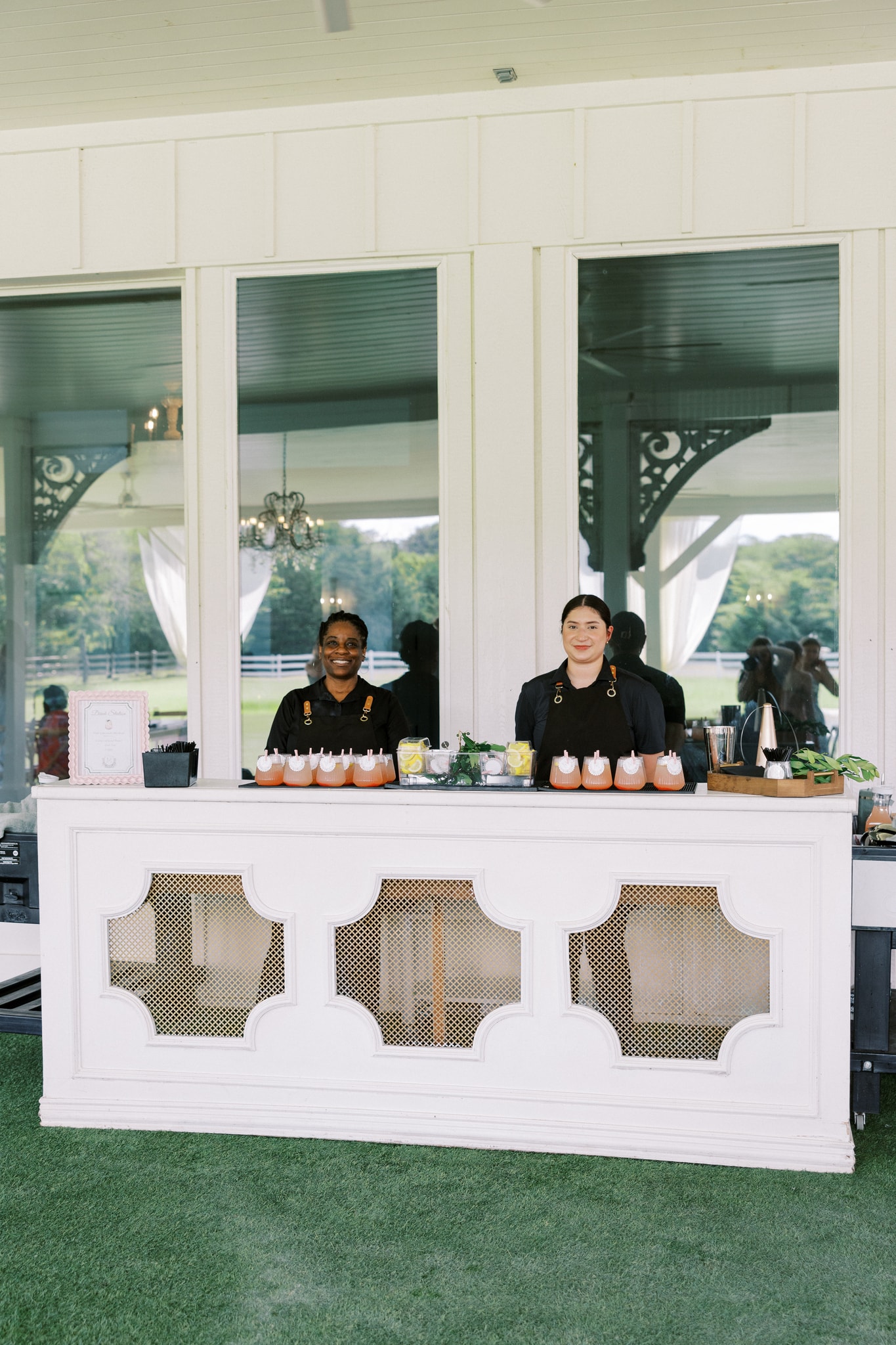 The French Farmhouse outdoor mobile bar — a white paneled bar with two bartenders in black uniforms, signature pink cocktails lined up, lemon and mint garnishes, set on the green lawn under the covered pavilion