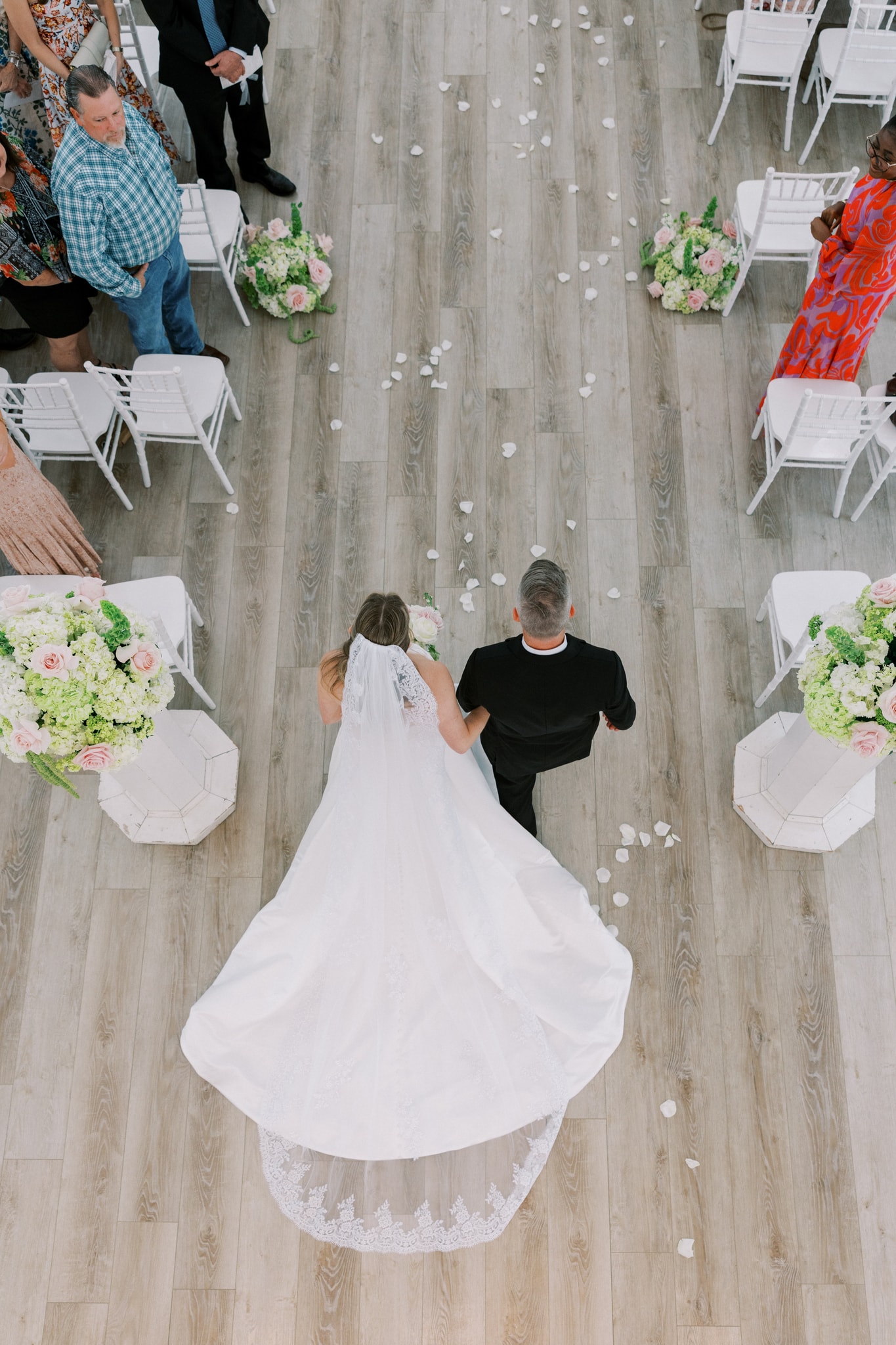 Aerial view of Morgan walking down the chapel aisle on her father's arm, her cathedral veil and lace-trimmed train spread wide, rose petals on the floor, guests standing on both sides