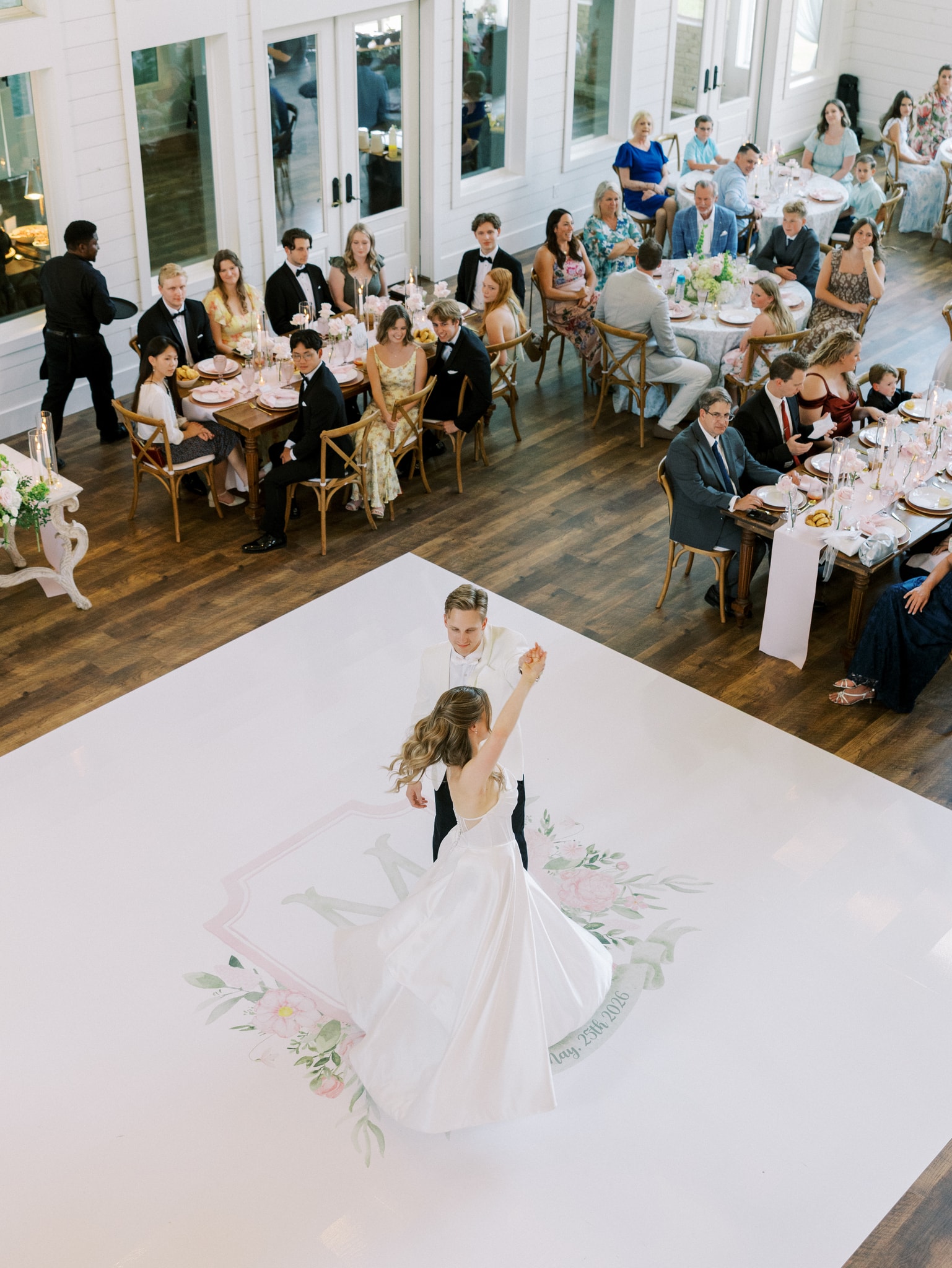 Aerial view of Morgan and Jack's first dance on their custom white dance floor printed with a watercolor floral crest and 'May 25th 2026', guests seated at farm tables around them