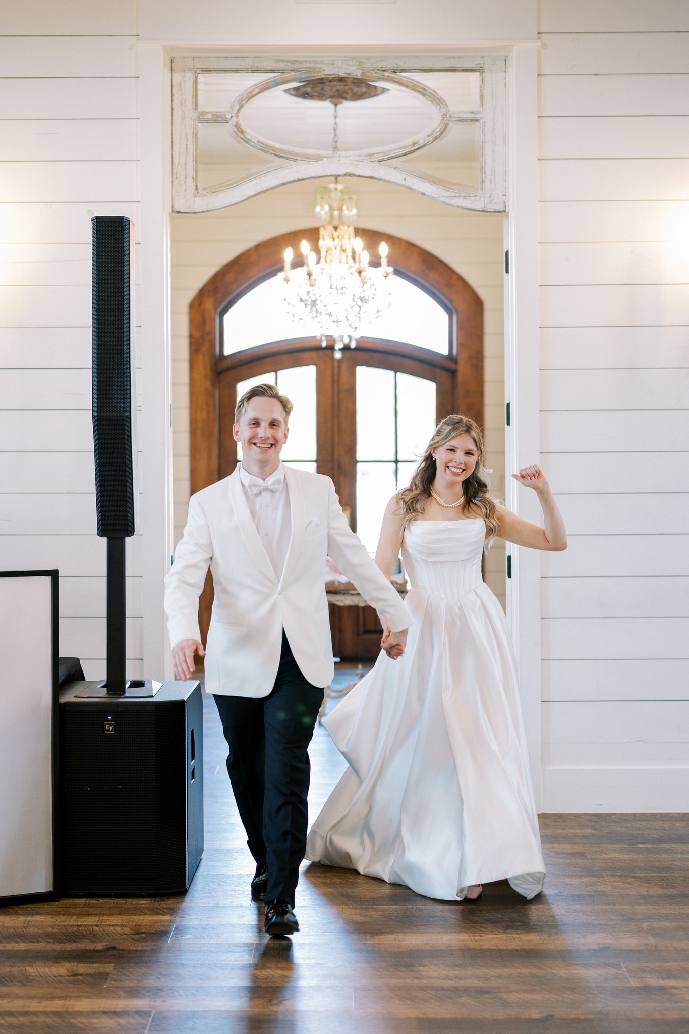 Morgan and Jack beaming as they make their grand entrance into the French Farmhouse reception hall, Jack in a white dinner jacket and Morgan in her strapless gown, the arched wood doors and crystal chandelier framing them from behind