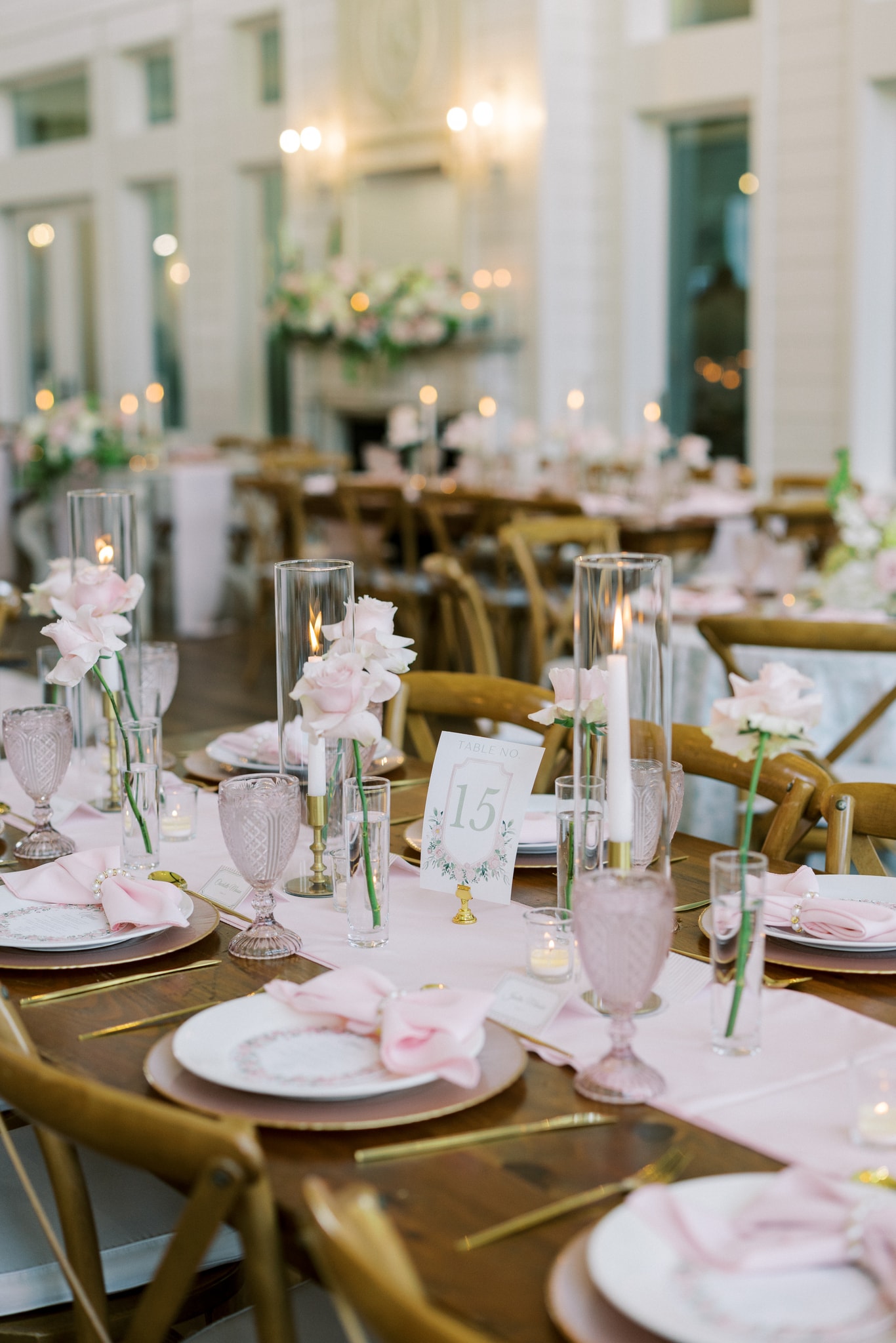 Morgan and Jack's reception tablescape — blush pink napkins, gold chargers, pink goblets, single stem blush roses in bud vases, taper candles in hurricane glasses, and a watercolor floral table number card, the full candlelit reception hall glowing behind