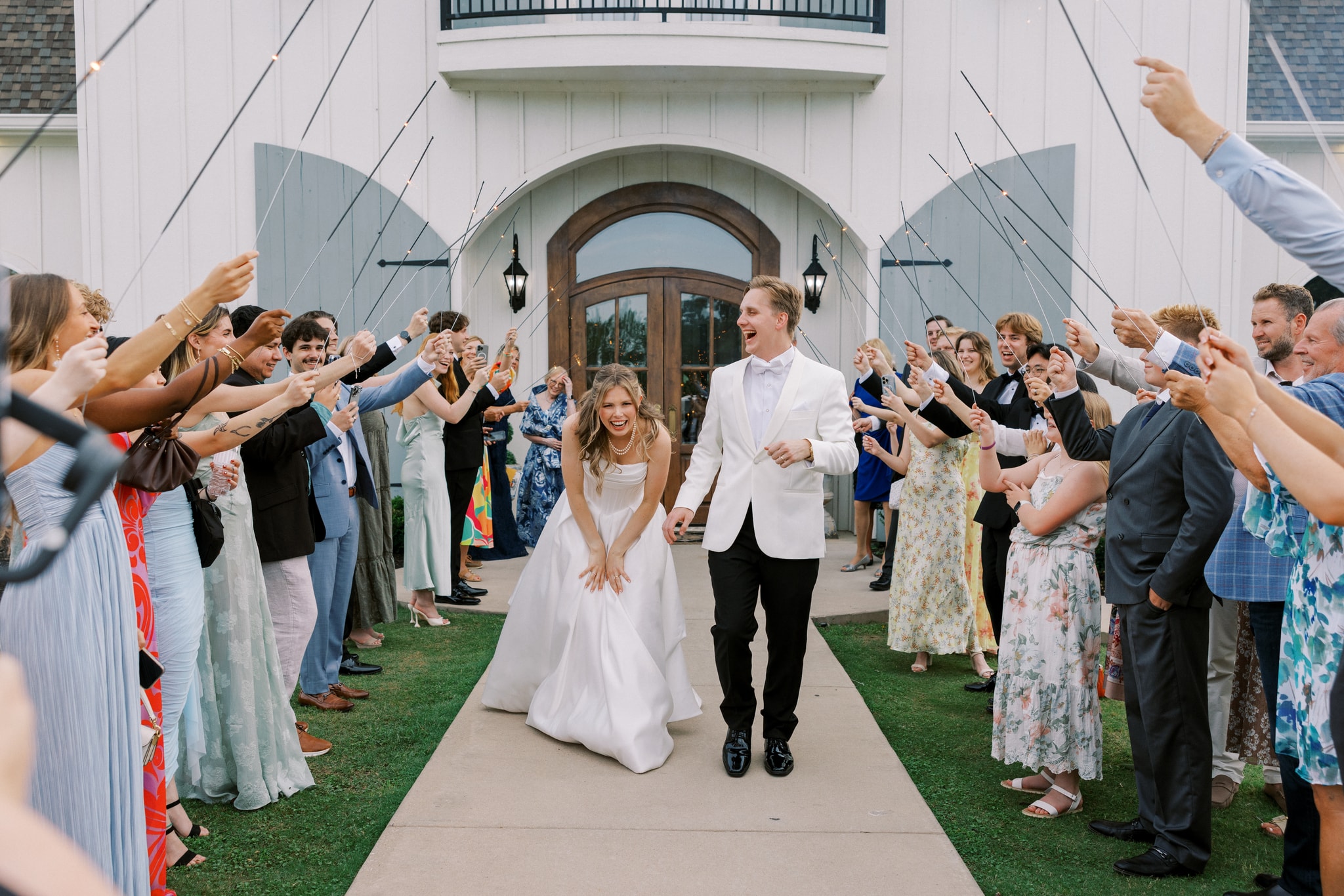 Morgan and Jack laughing and running through a tunnel of sparklers held by their guests on the front lawn of The French Farmhouse Venue, the white board-and-batten facade and arched wood doors glowing behind them at dusk