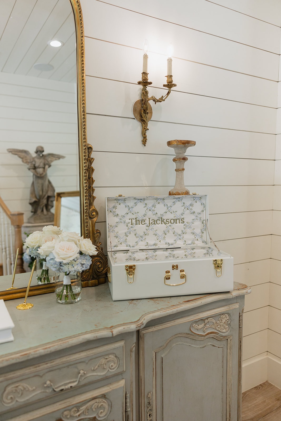 Custom 'The Jacksons' card box on the antique French dresser in the chapel foyer at The French Farmhouse Venue, with white roses, blue hydrangeas, gold mirror, and brass wall sconce