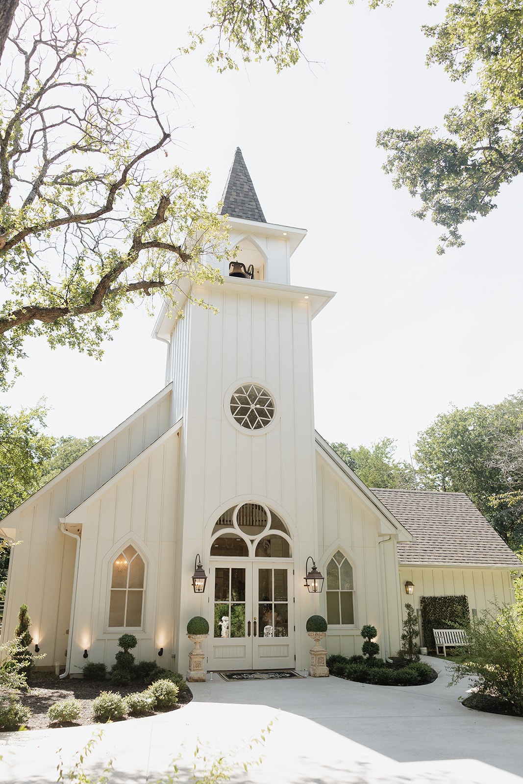The French Farmhouse chapel exterior on Nina and Gage's 4th of July wedding day — white steeple, arched doors, topiary urns, and summer trees