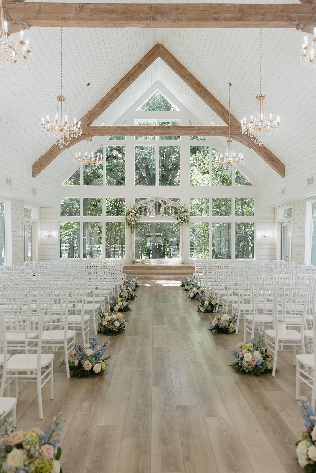 Wide interior shot of The French Farmhouse chapel set up for Nina and Gage's ceremony — white chiavari chairs, blue and blush aisle florals by JG Floral Design, antique arch, crystal chandeliers, and floor-to-ceiling windows