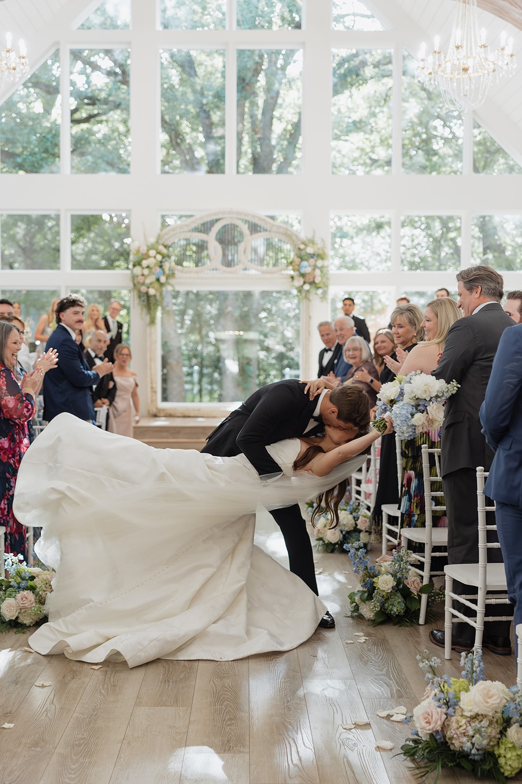 Gage dipping Nina into their first kiss during the recessional at The French Farmhouse chapel — antique arch, floor-to-ceiling windows with summer trees, and the entire room on their feet cheering