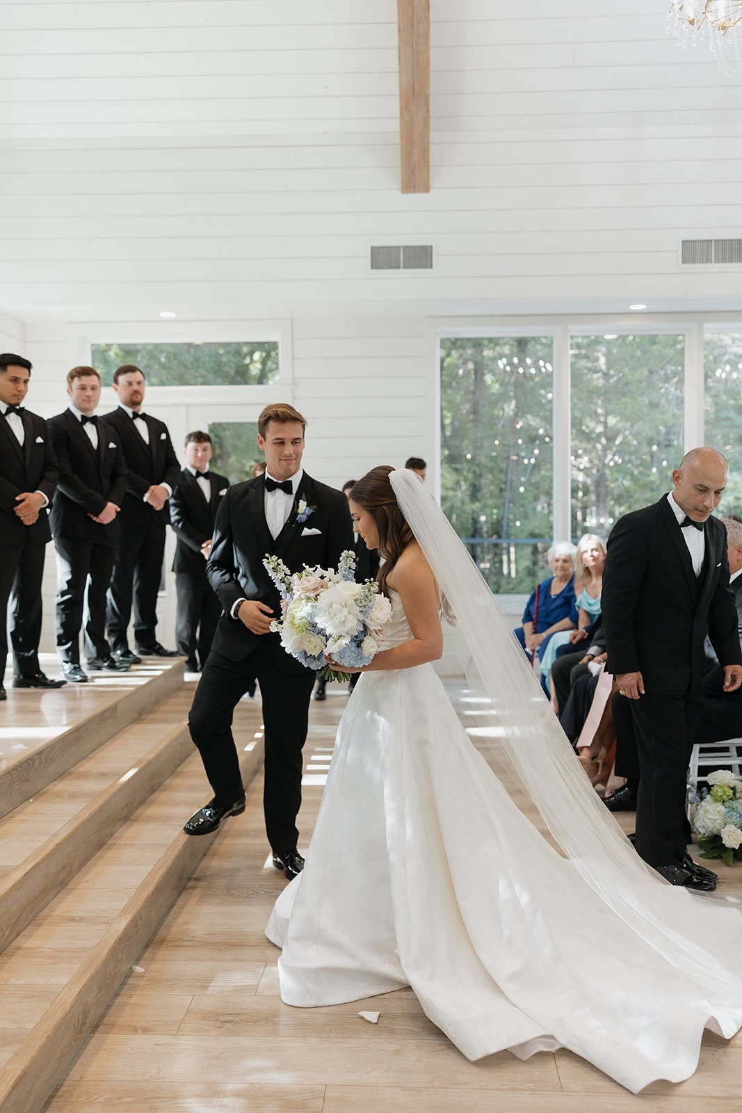 Gage stepping down from the altar to meet Nina at the chapel steps, groomsmen lined up behind him, Nina looking down with her bouquet and cathedral veil flowing