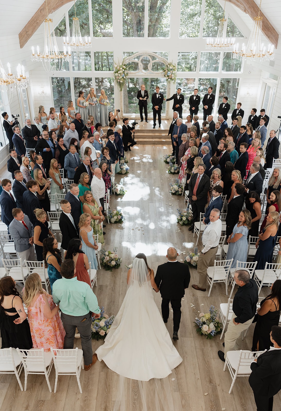 Breathtaking aerial view of Nina walking down the aisle with her dad at The French Farmhouse chapel — the entire room on their feet, Gage and groomsmen waiting at the antique arch, blue and blush aisle florals lining every row