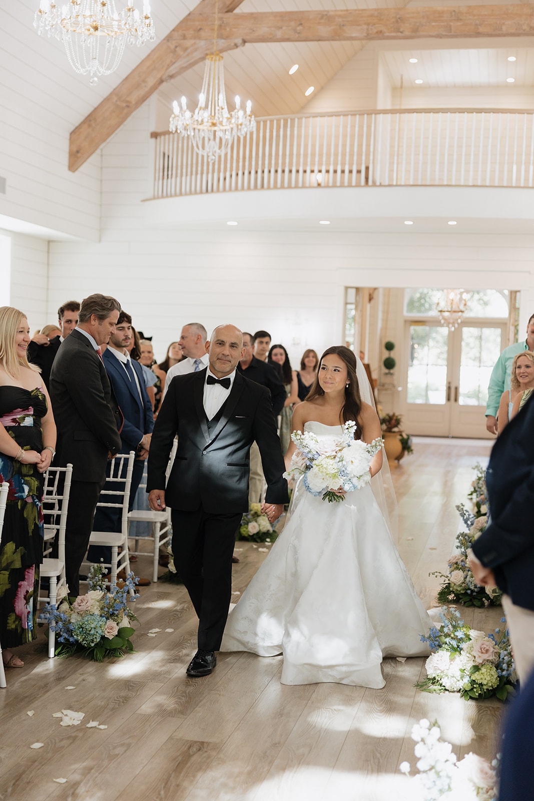 Nina walking down the aisle with her dad at The French Farmhouse chapel — crystal chandeliers overhead, blue and blush aisle florals, guests standing and smiling on both sides