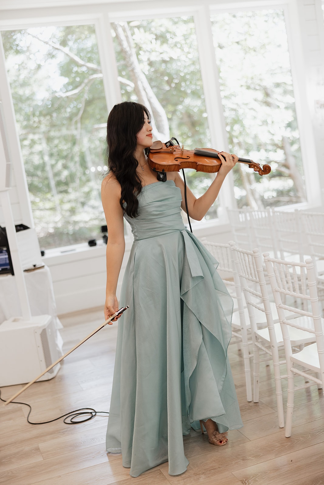 Faith Fang of Faith Fang Music playing violin in a light blue gown inside The French Farmhouse chapel with floor-to-ceiling windows and summer trees behind her