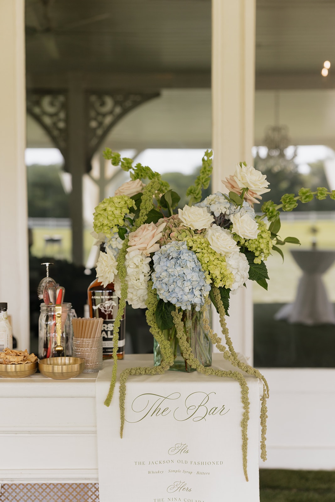 The bar setup at Nina and Gage's cocktail hour under the covered patio at The French Farmhouse Venue — JG Floral Design arrangement of blue hydrangeas, blush roses, and green amaranthus with 'The Bar' sign listing The Jackson Old Fashioned and The Nina Colada