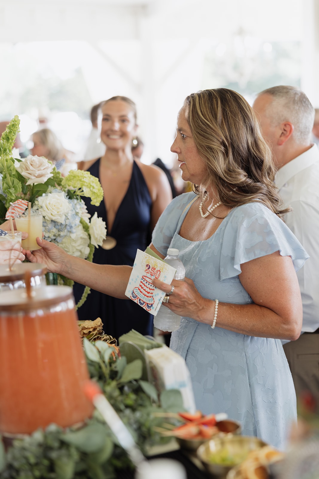 Guests mingling and grabbing drinks at the bar under the covered patio during cocktail hour at The French Farmhouse Venue — blue hydrangea and white rose florals, patriotic drink stirrers, and the warm summer afternoon light