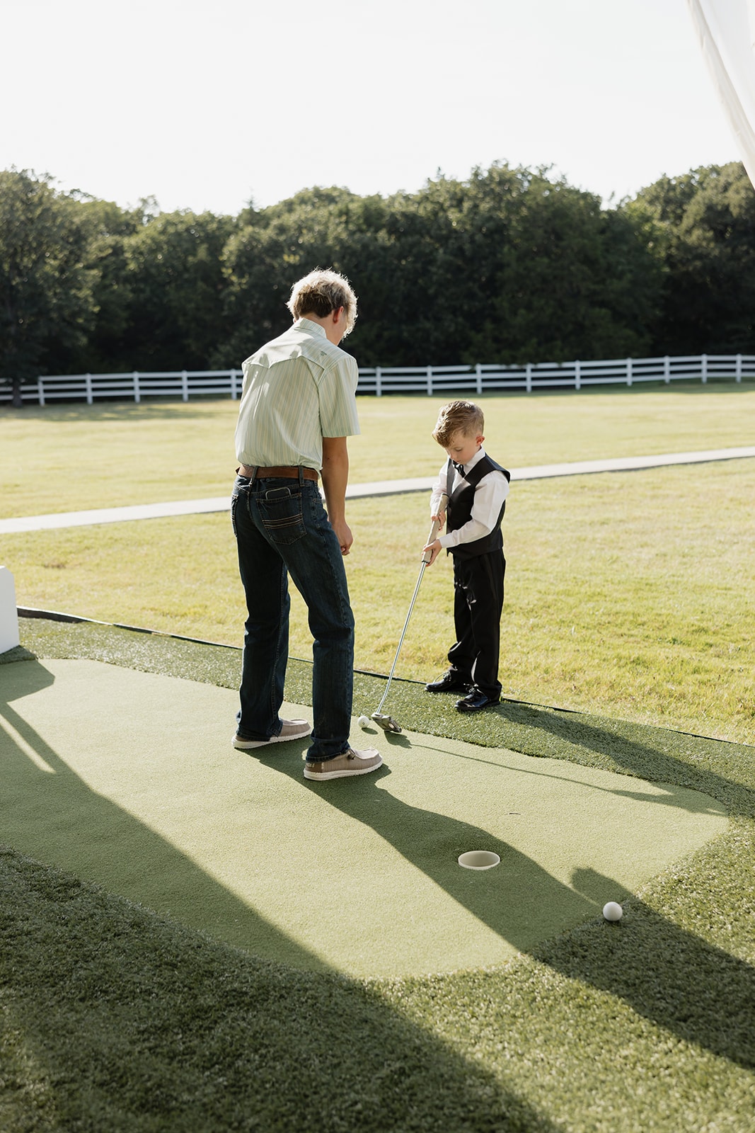 Two young guests in their wedding best playing on the putting green during cocktail hour at The French Farmhouse Venue — white fence and summer trees stretching out behind them in golden afternoon light