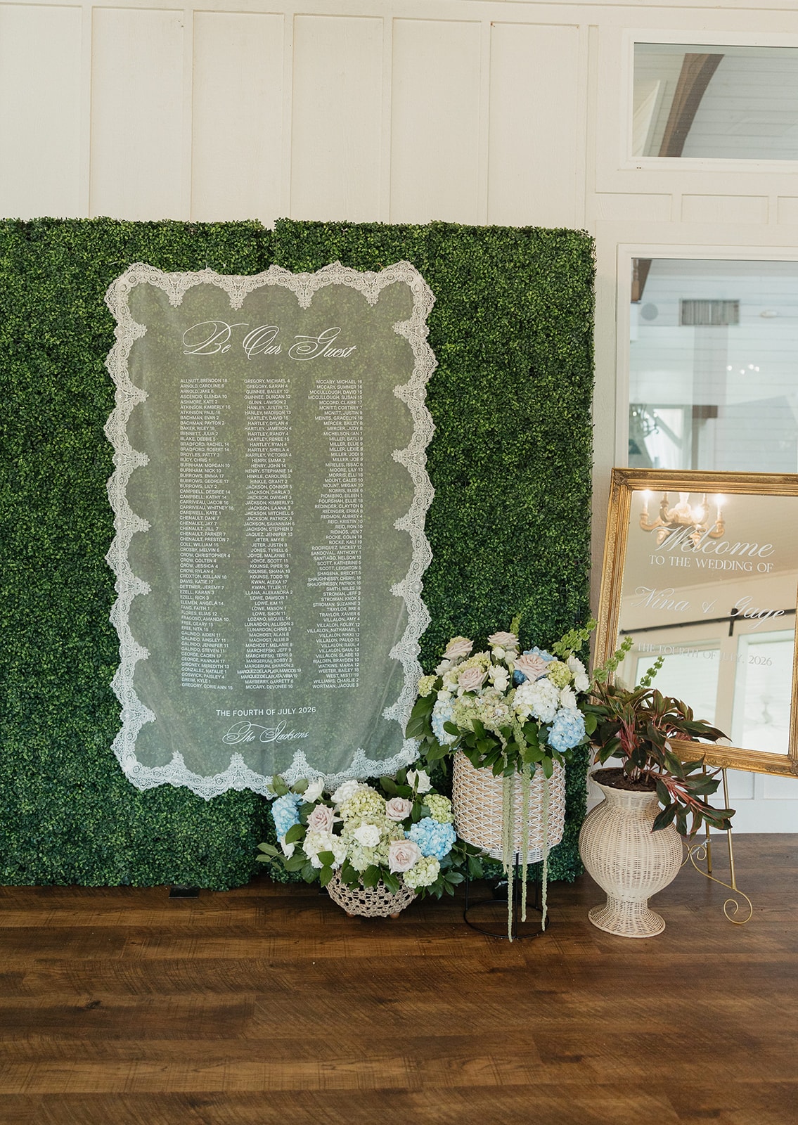 'Be Our Guest' acrylic seating chart with lace border displayed against the boxwood hedge wall during cocktail hour — The Jacksons, The Fourth of July 2026