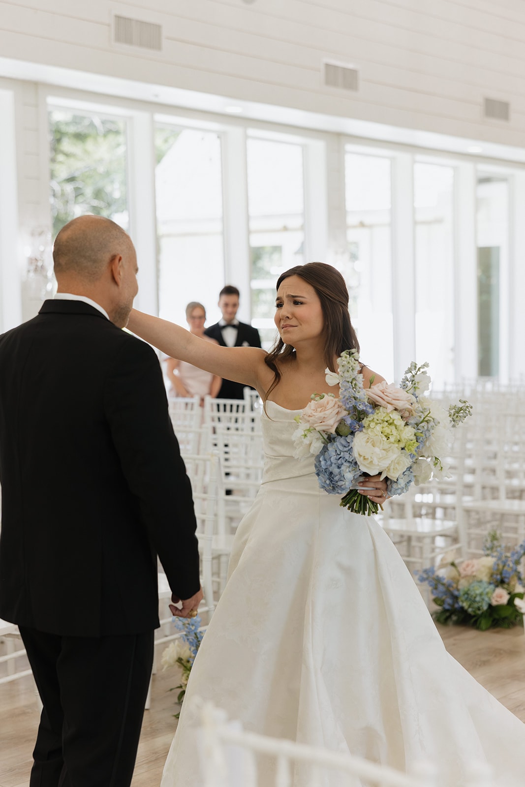 Nina reaching out to tap her dad's shoulder for their first look inside The French Farmhouse chapel, holding her blue hydrangea and blush rose bouquet